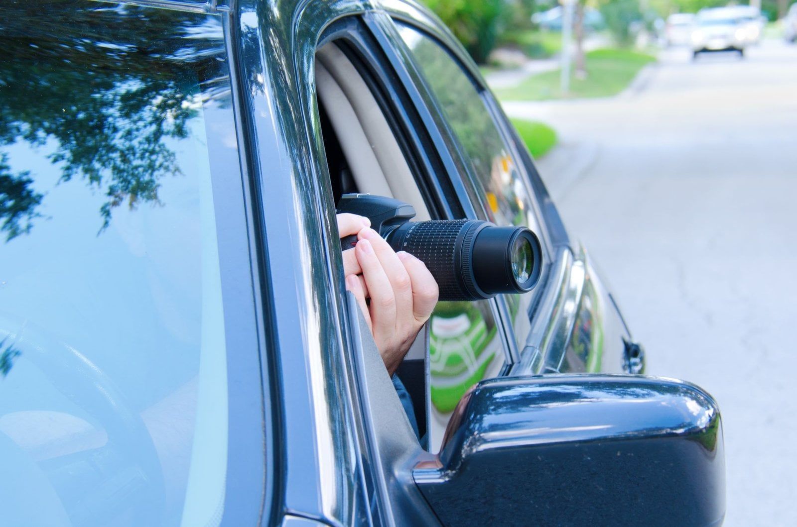 A person inside a car holding a camera lens out the open window, angled toward the street.