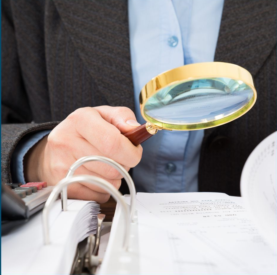 A person in a business suit uses a magnifying glass to inspect documents in an open binder.