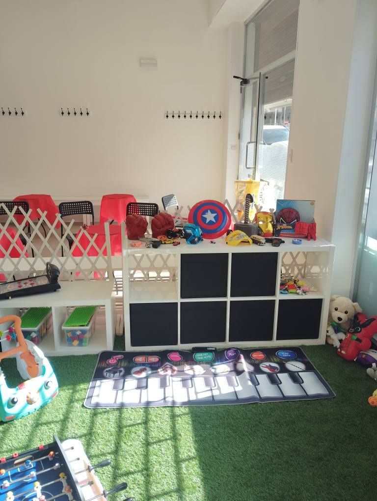Sala giochi con giocattoli su mensole e un tappeto musicale. Sono presenti anche sedie rosse e un biliardino.