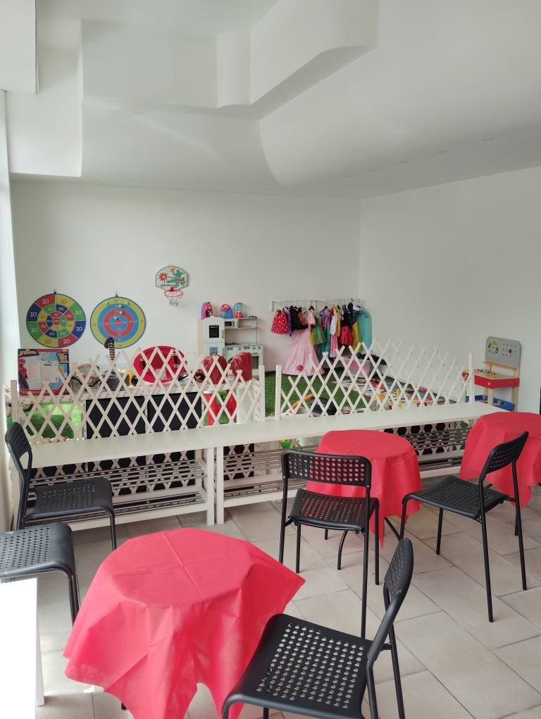 Una stanza con tavoli rossi rotondi, sedie nere e una staccionata bianca decorata con oggetti; pareti per lo più bianche.