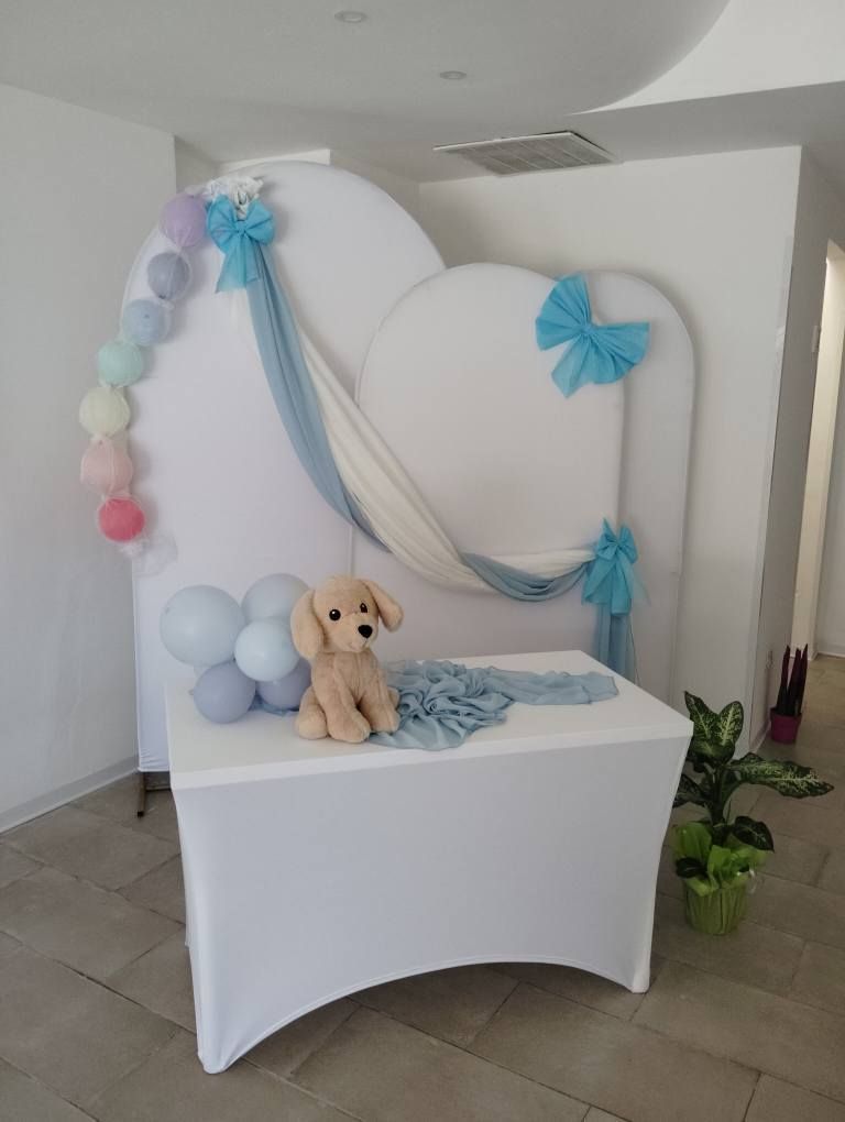 Tavolo apparecchiato di bianco con un cucciolo di peluche, palloncini e decorazioni color pastello per una festa.
