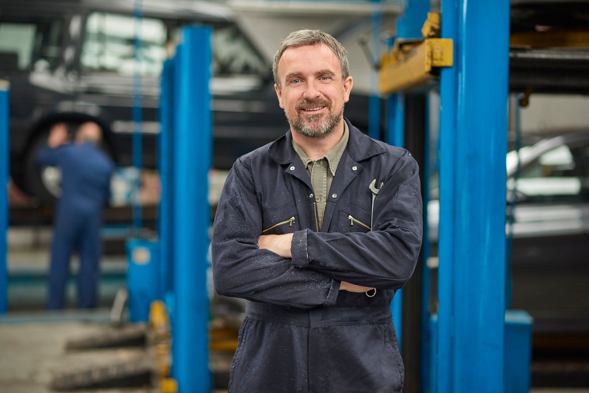 A smiling garage owner stands looking to camera from his busy auto repair shop
