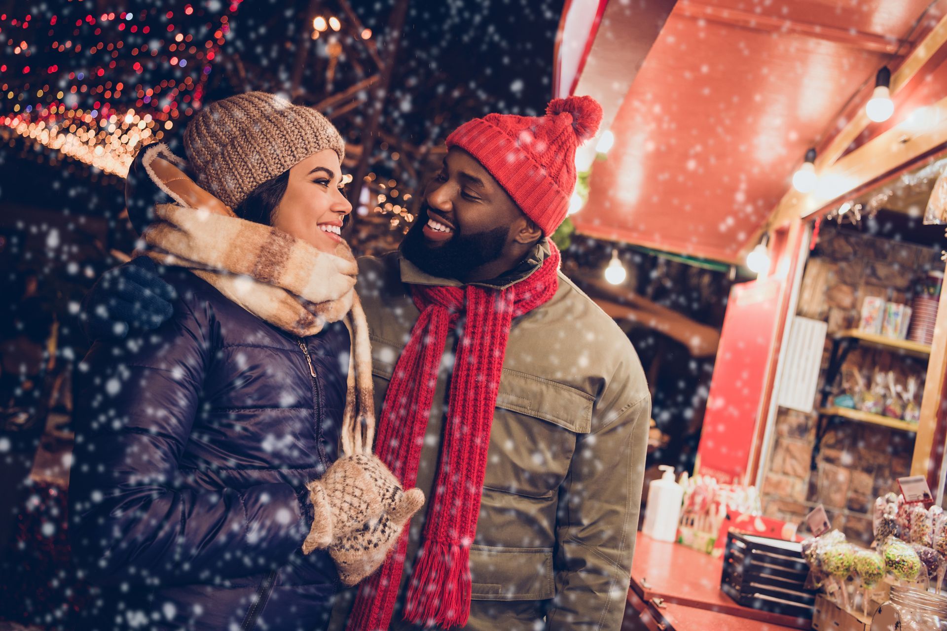 Couple smiles at each other at an outdoor market in the snow, wearing winter hats and scarves.