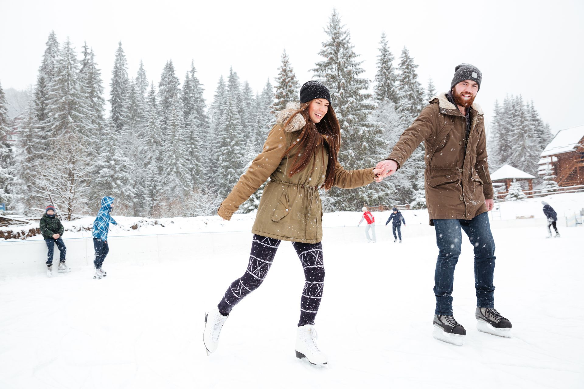 Couple ice skating, holding hands. Snowy scene, winter clothing. Others skate in background.