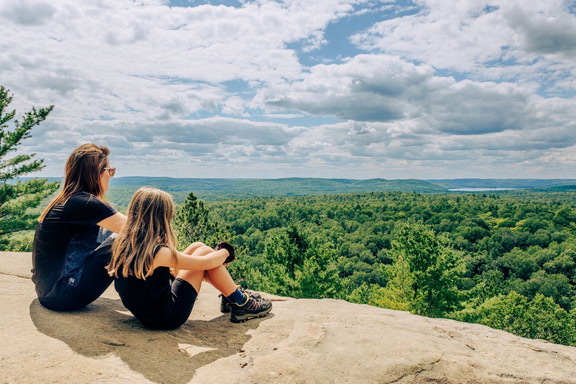 A woman and a child are sitting on a rock overlooking a forest.