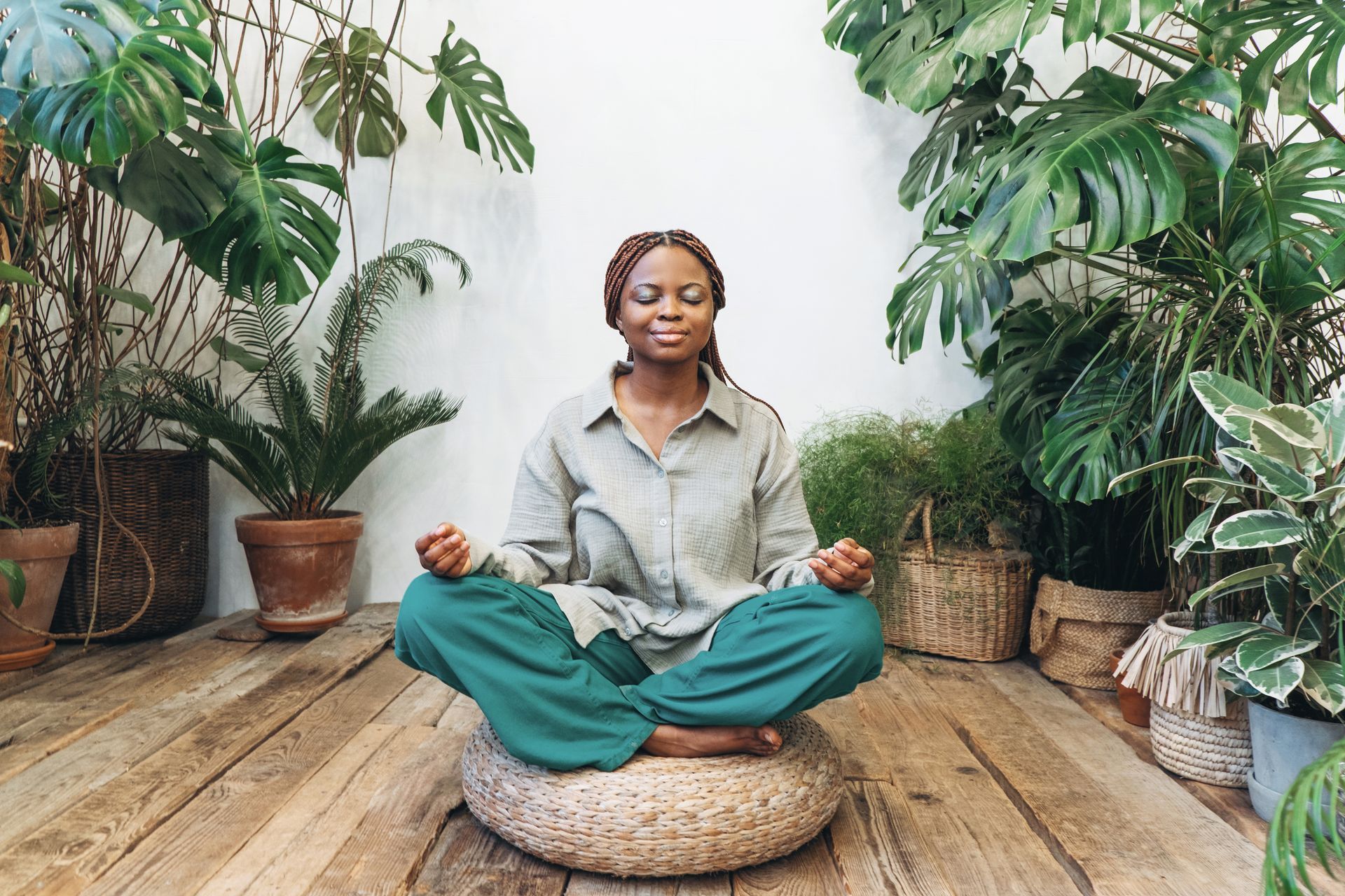 A woman is sitting in a lotus position on a wooden floor surrounded by plants.