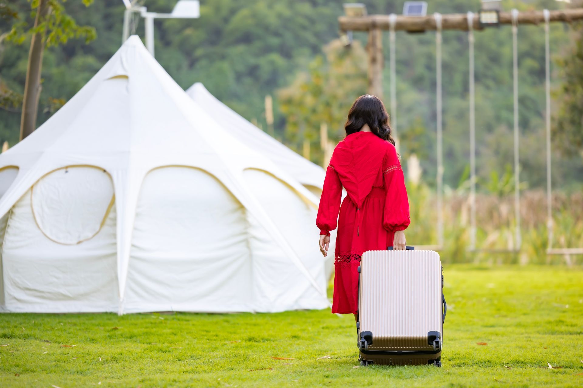 Woman in red dress pulling a suitcase on a grassy lawn near a white tent.