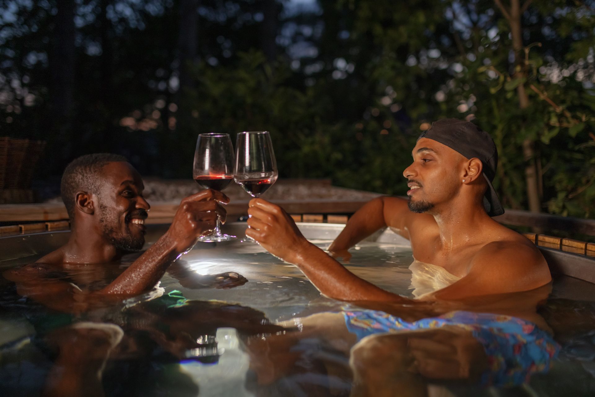 Two men toasting wine glasses in a hot tub at night. One wears a cap. Both are smiling.