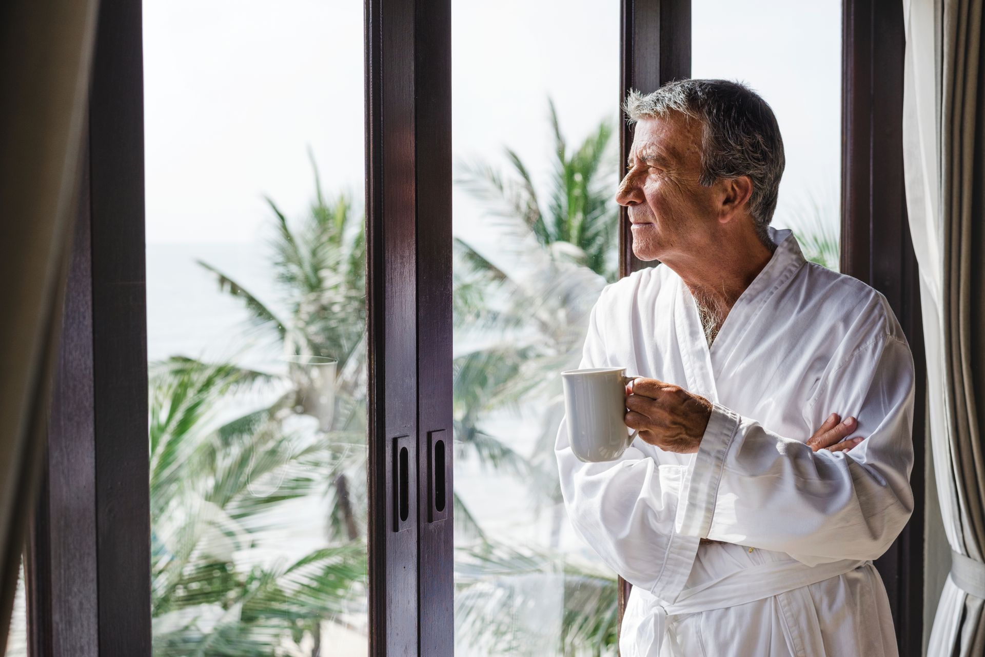 Older man in white robe, holding mug, looking out window with tropical view.