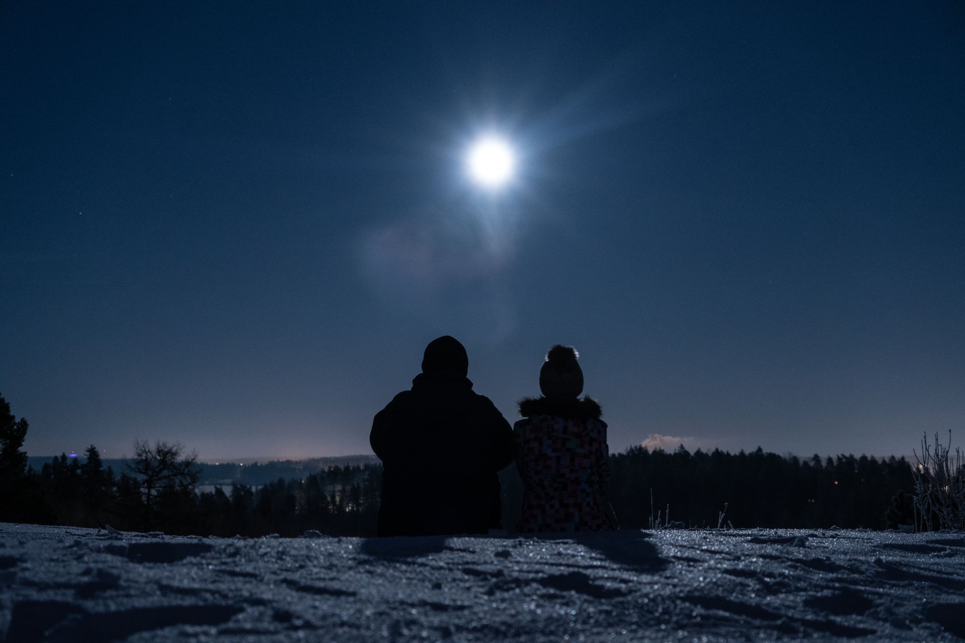 A man and a woman are sitting on top of a snow covered hill looking at the moon.