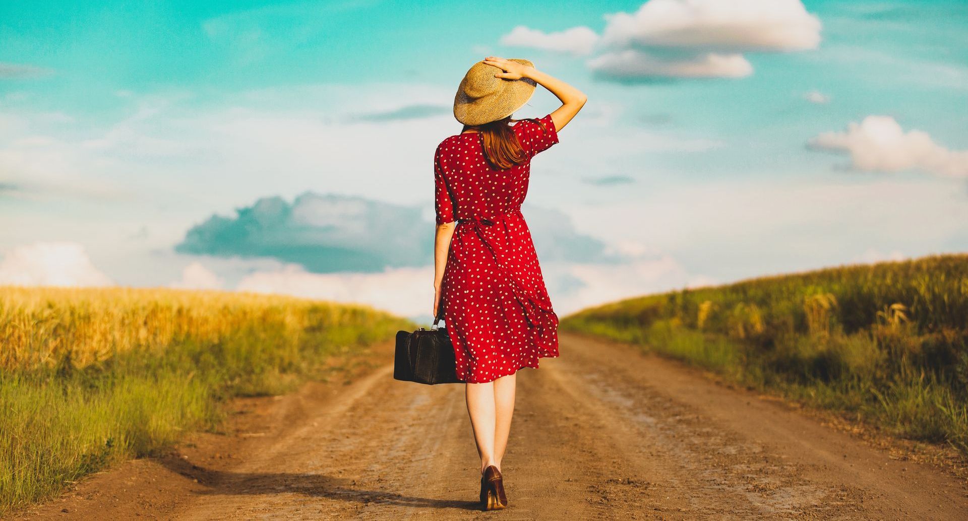 A woman in a red dress and straw hat is walking down a dirt road.