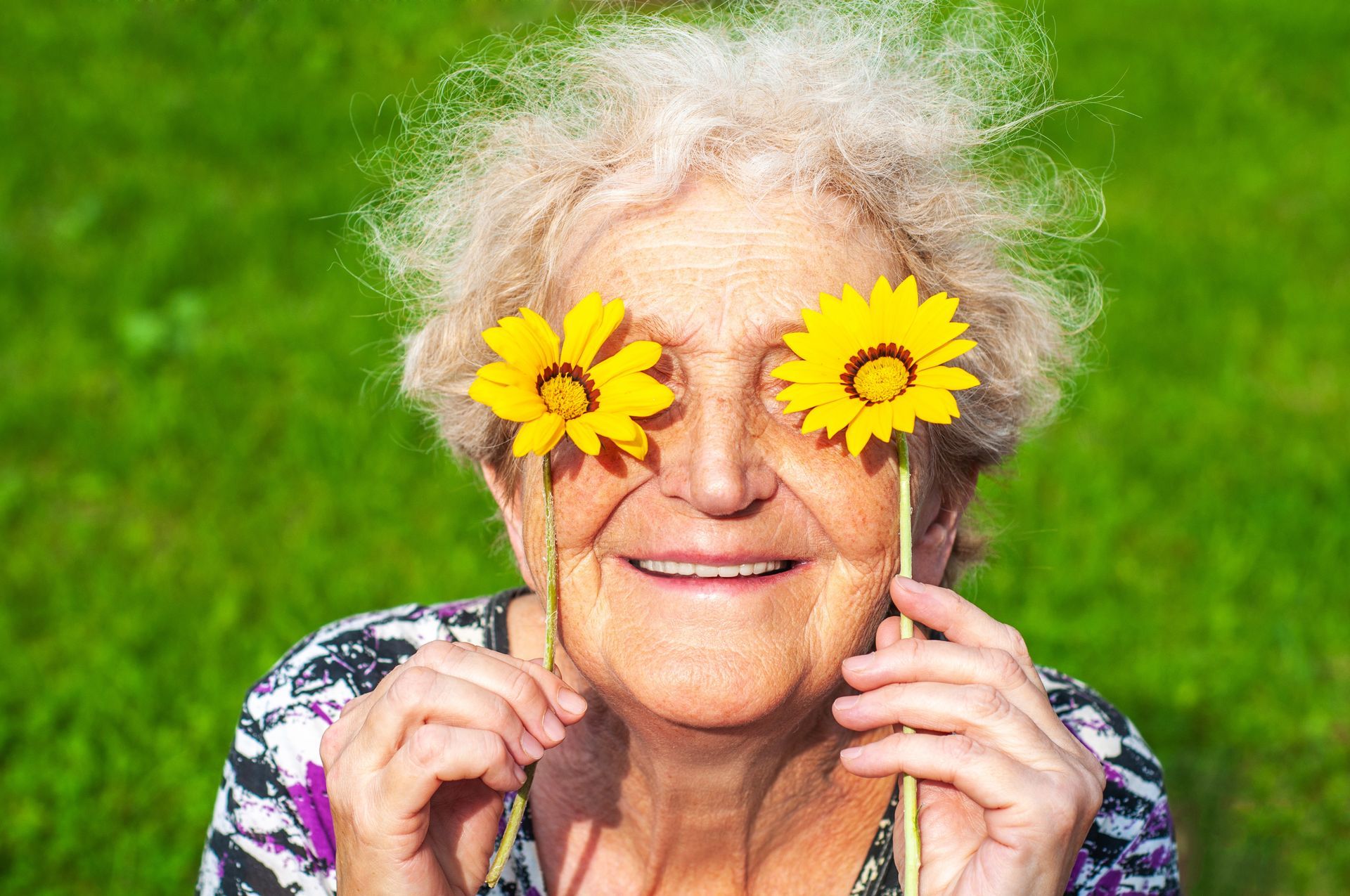 An elderly woman is holding two yellow flowers in front of her eyes.