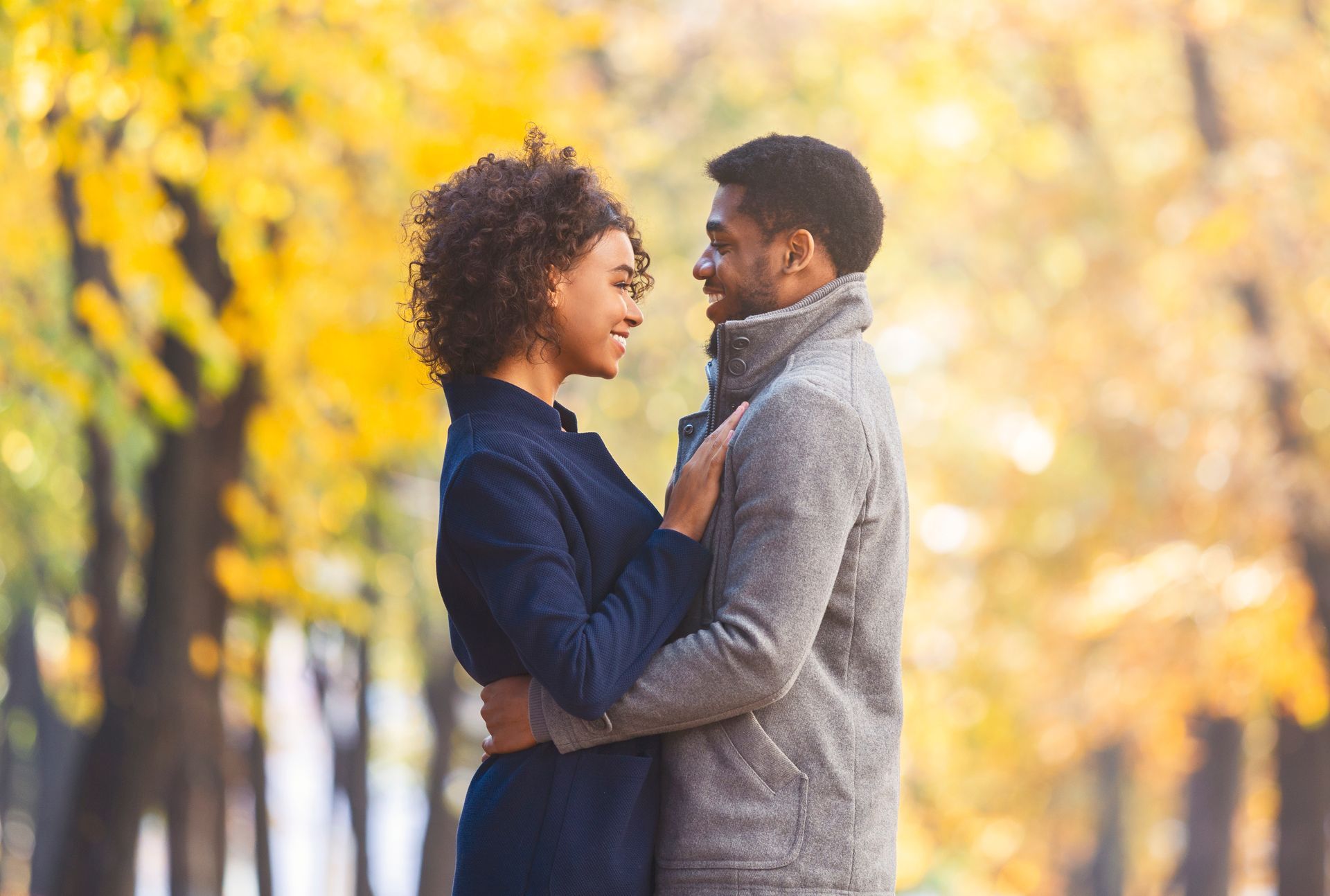 Couple embracing, smiling, and looking at each other in a park with yellow fall foliage.