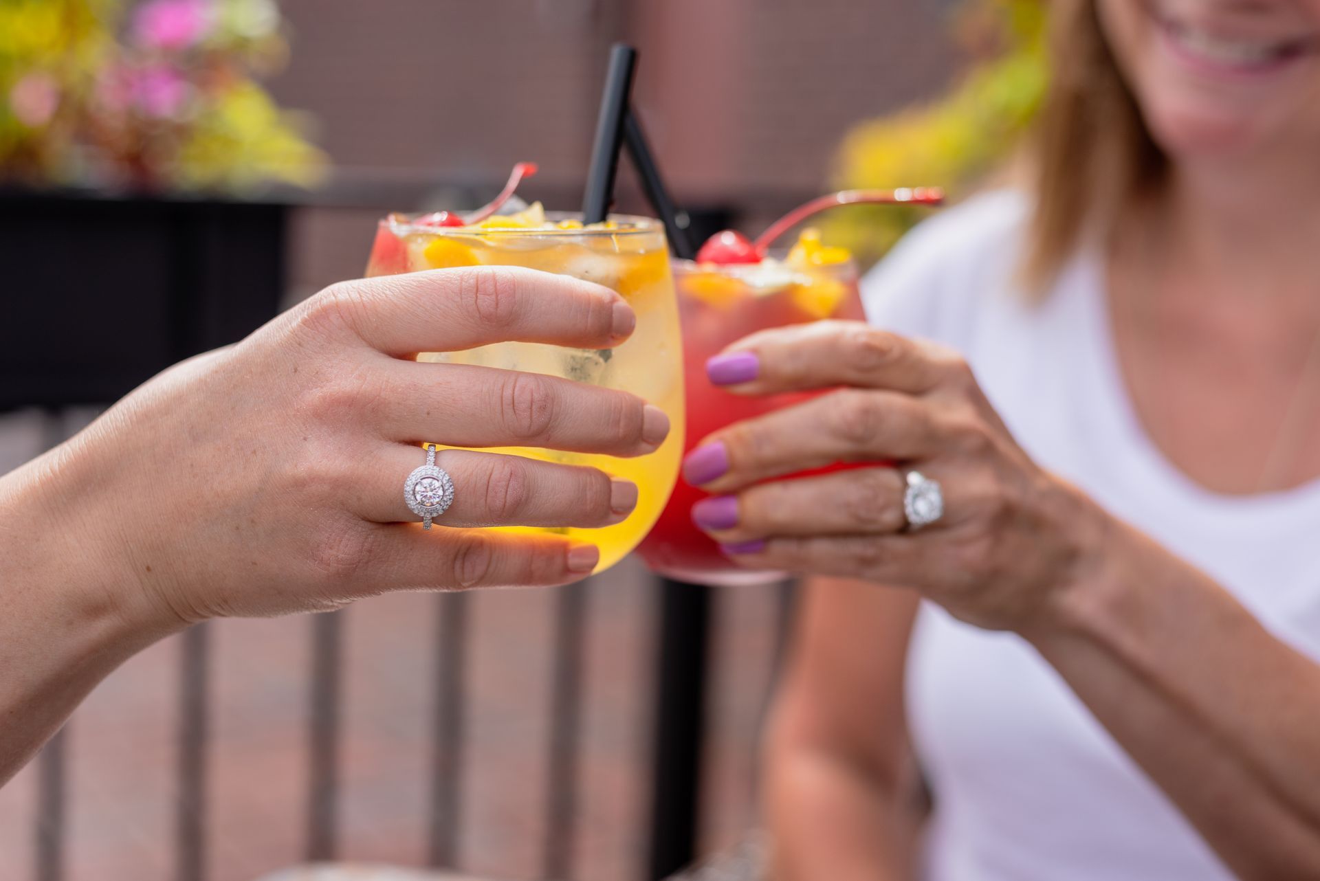 Two women toasting with cocktails, one yellow, one red, on a patio, showing off engagement rings.