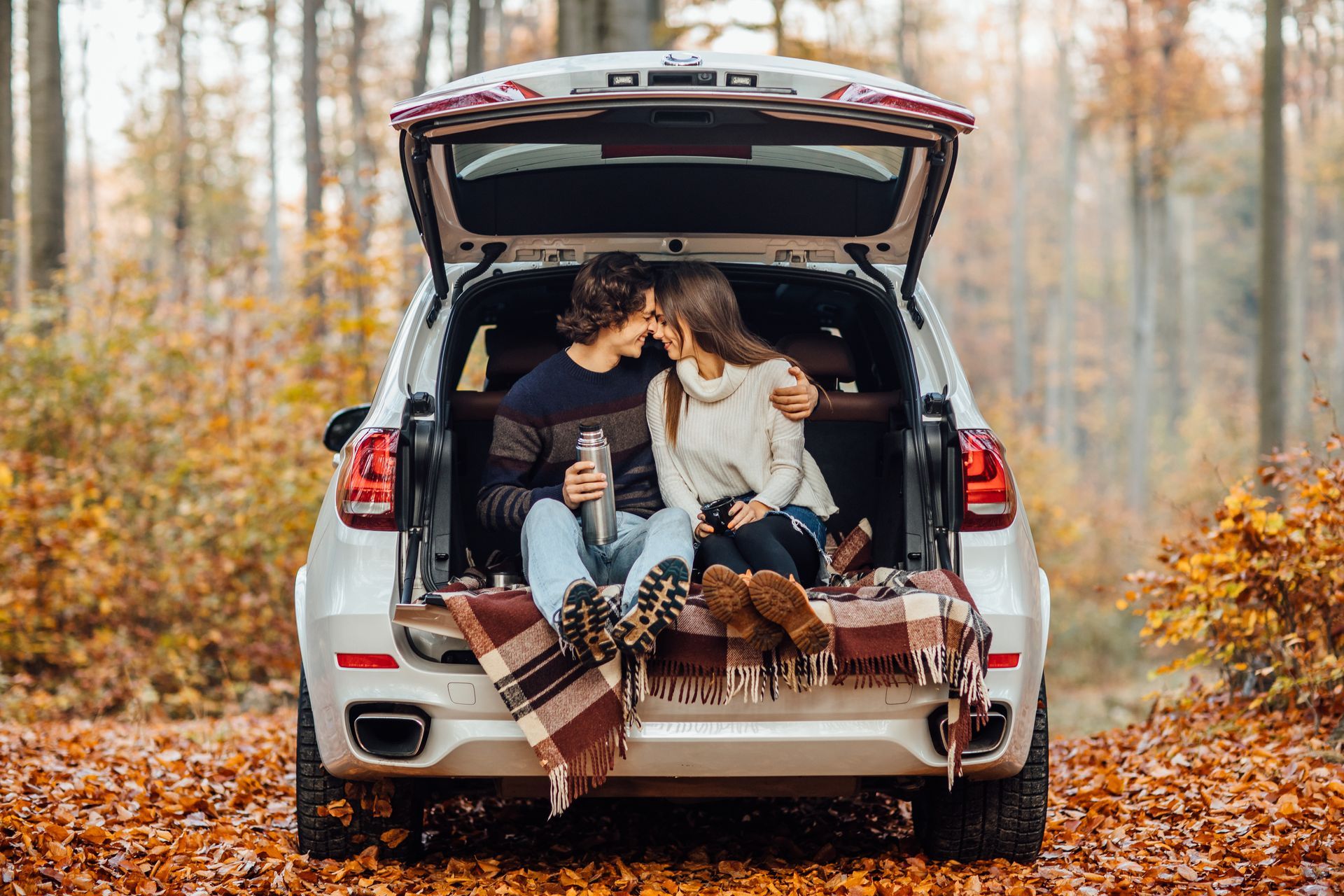 Couple kissing in open SUV trunk, fall setting, with blanket, trees, and leaves.