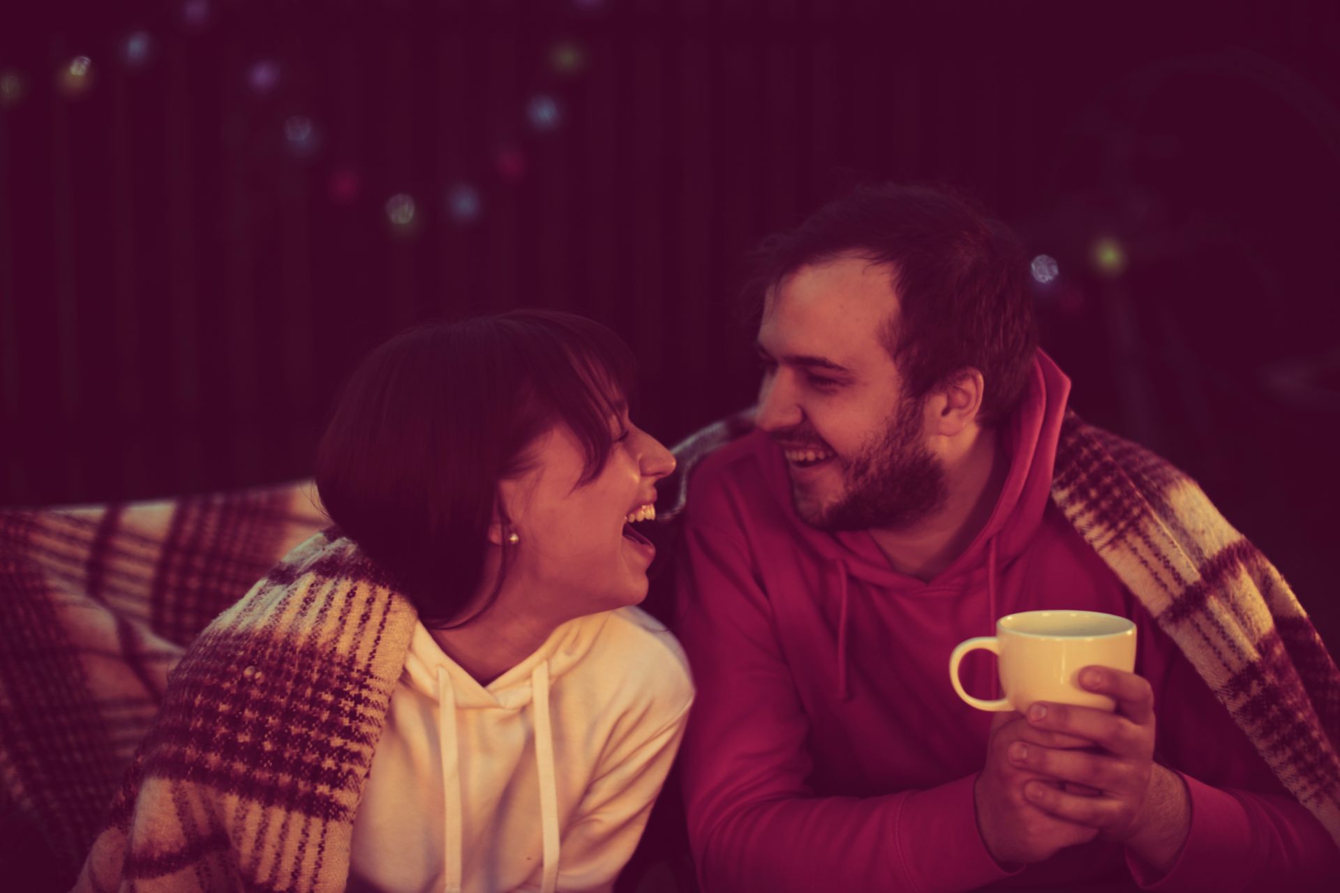 Couple laughing, wrapped in a blanket, holding a mug, under string lights.