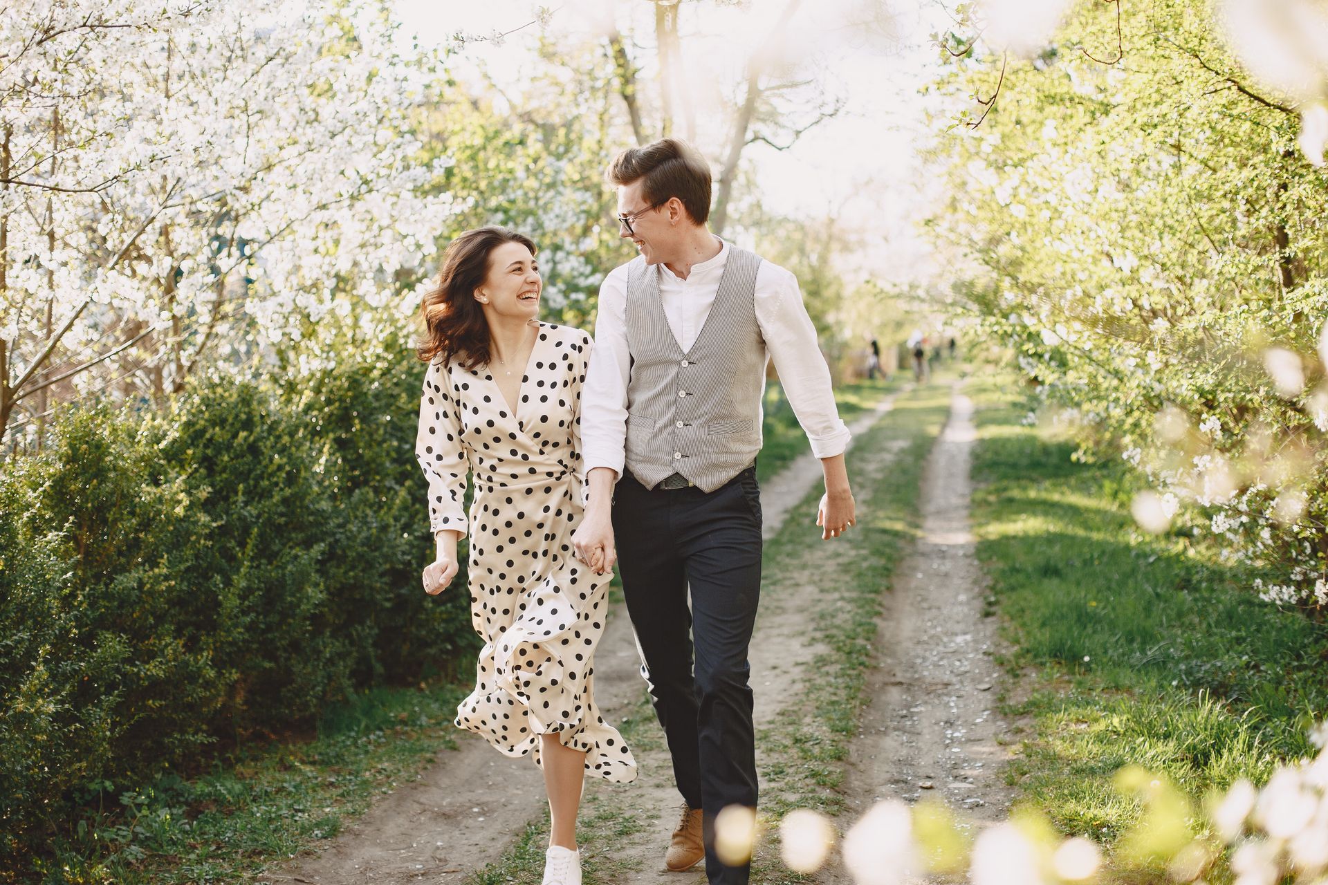 Couple walking hand-in-hand on a path lined with blooming trees; smiling, cheerful expressions; sunny day.