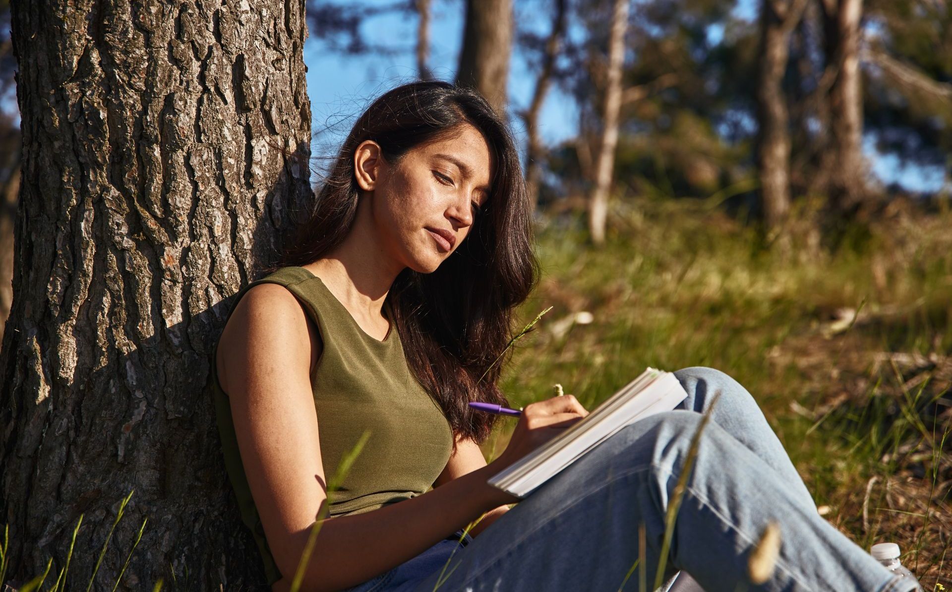 A woman is sitting under a tree in the woods writing in a notebook.