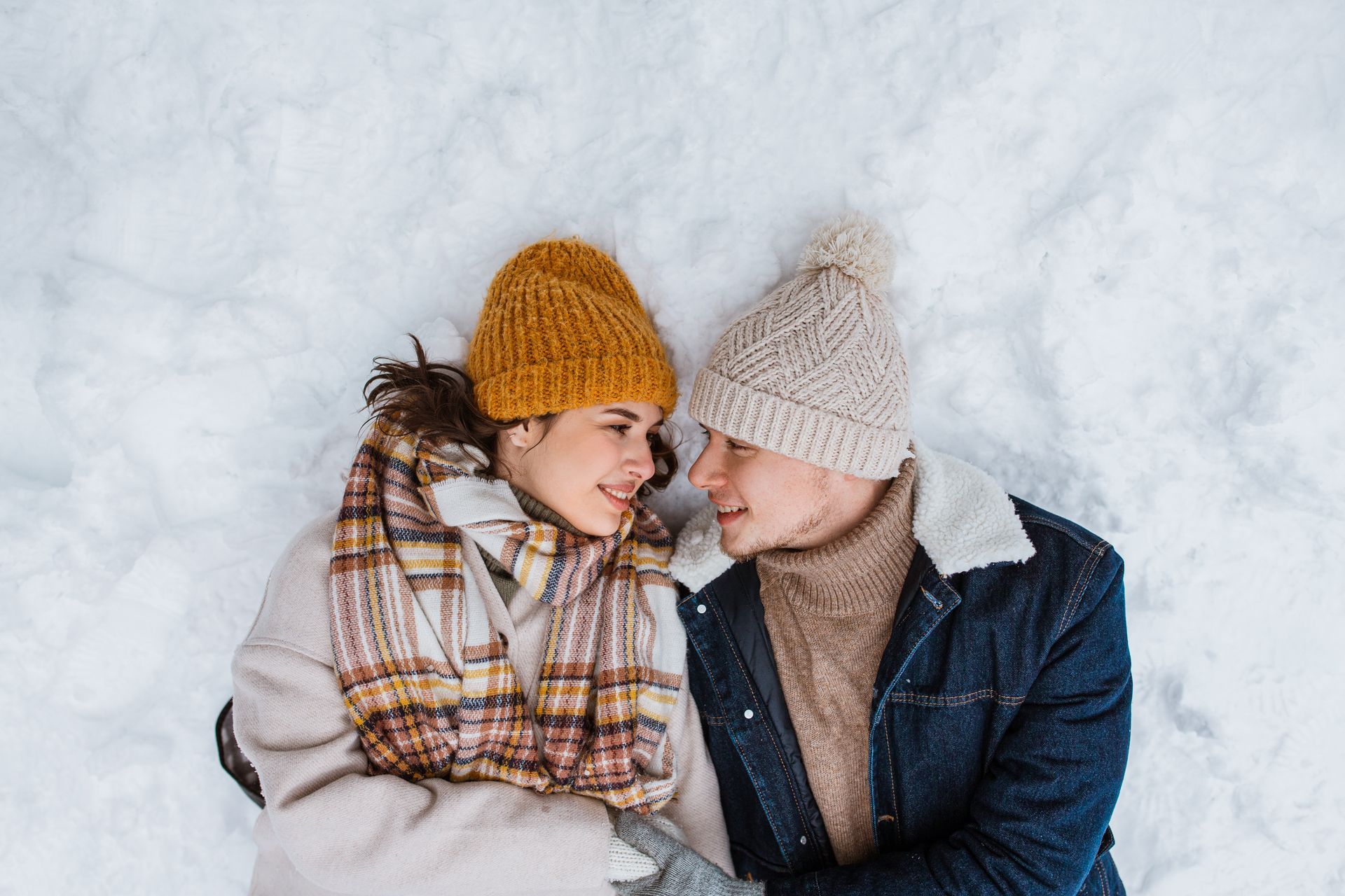 Couple lying in snow, gazing at each other. They wear winter hats, scarves, and jackets.
