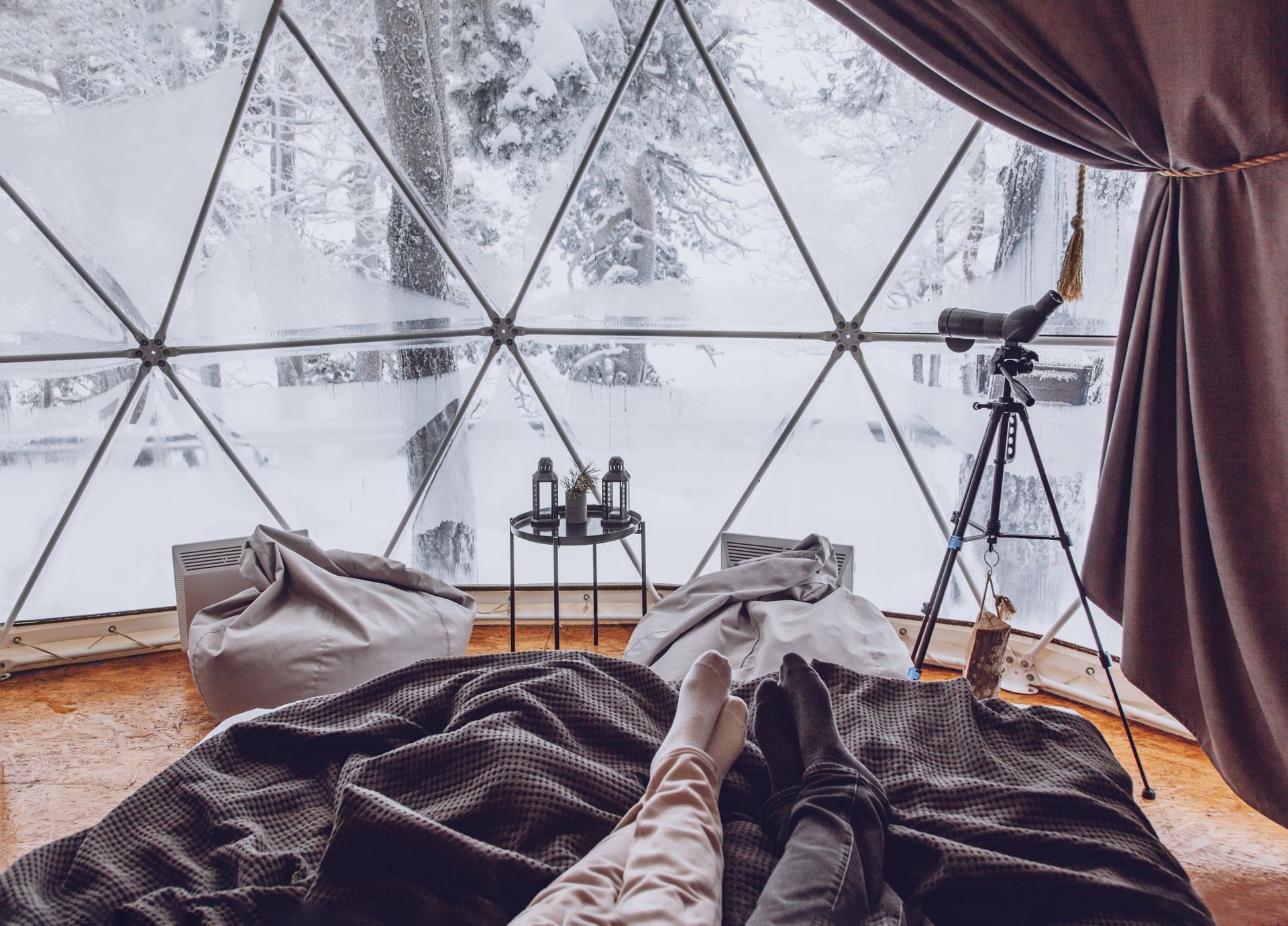 Inside a geodesic dome, feet rest on a bed looking out at a snowy landscape through triangular windows.