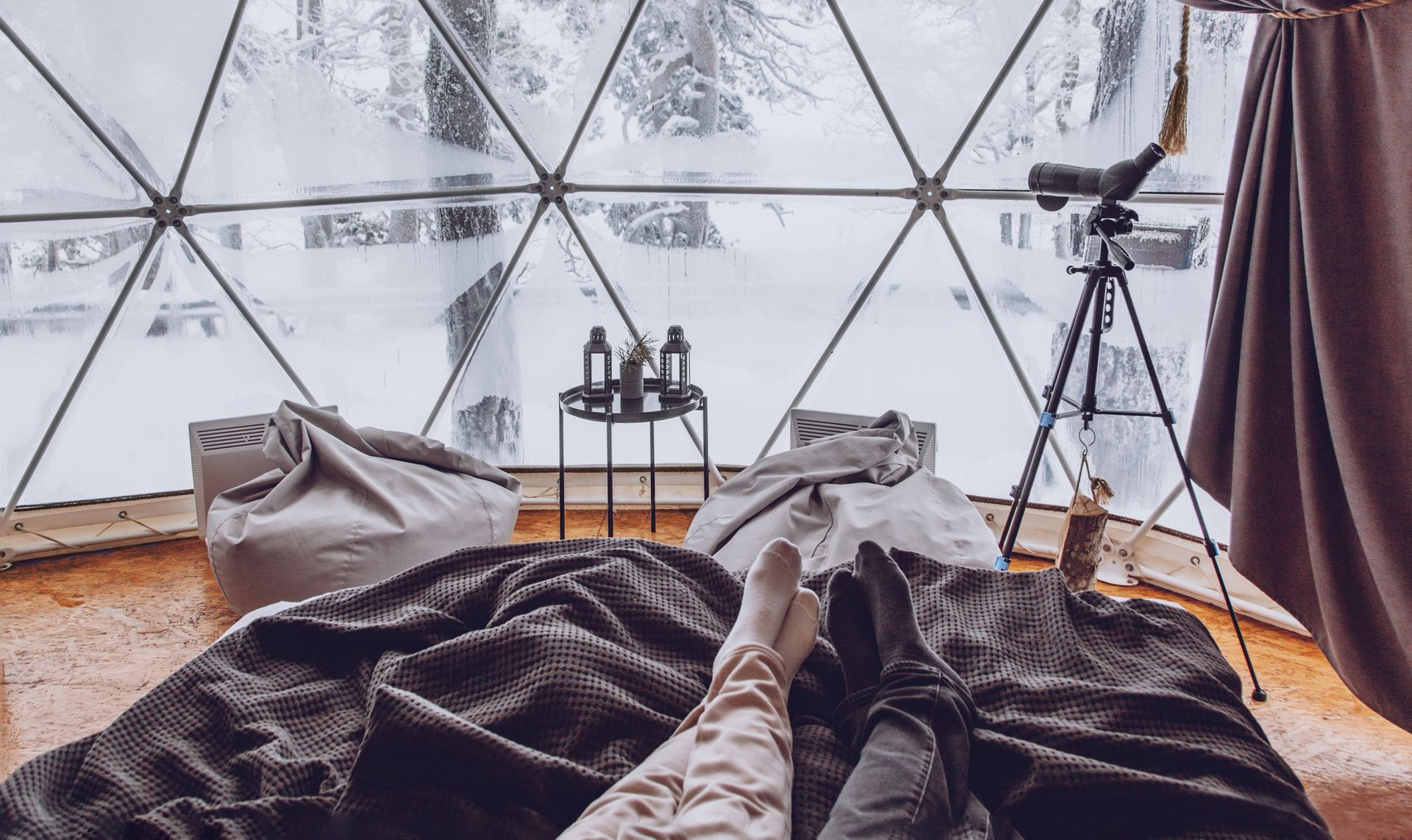 Couple relaxing in a dome with a snowy view. Beanbags, telescope, and blankets create a cozy setting.