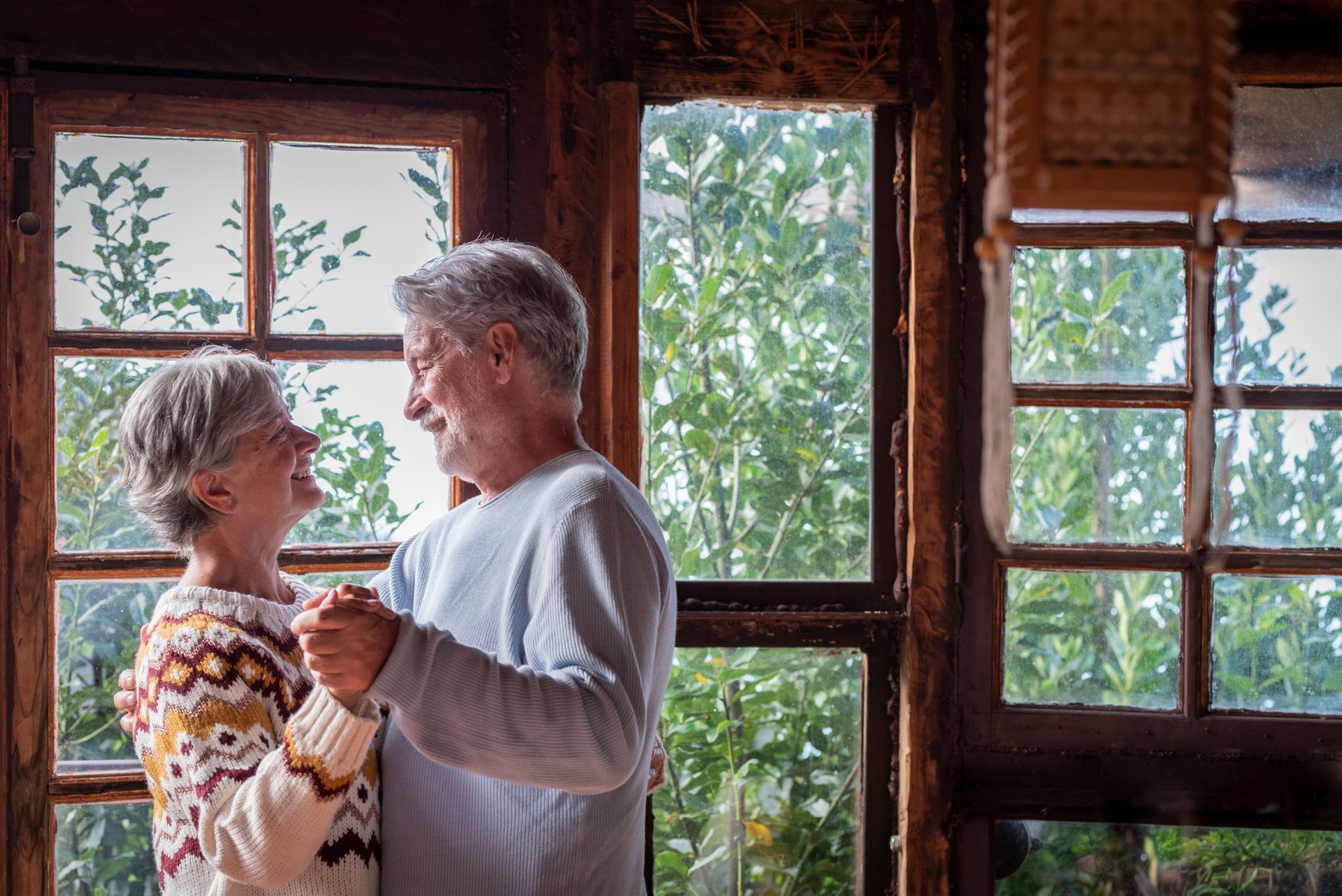 An elderly couple is dancing in front of a window.