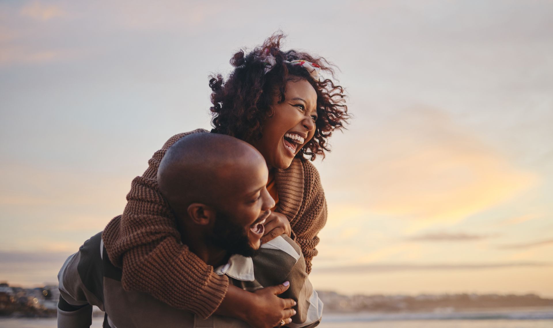 Couple laughing, man giving woman piggyback ride outdoors at sunset.