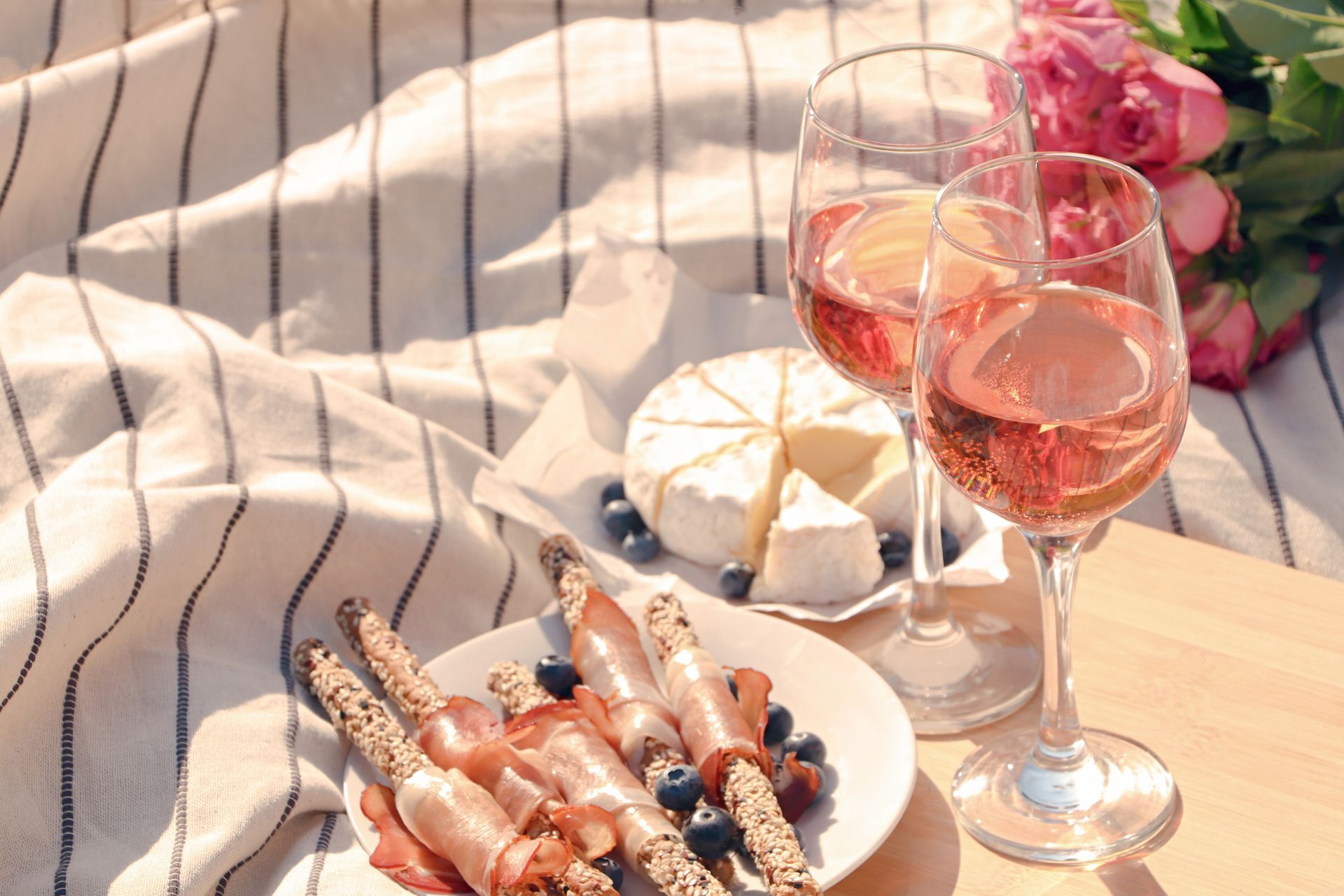 Picnic setup with wine, cheese, prosciutto-wrapped breadsticks, blueberries, and flowers on a striped blanket.