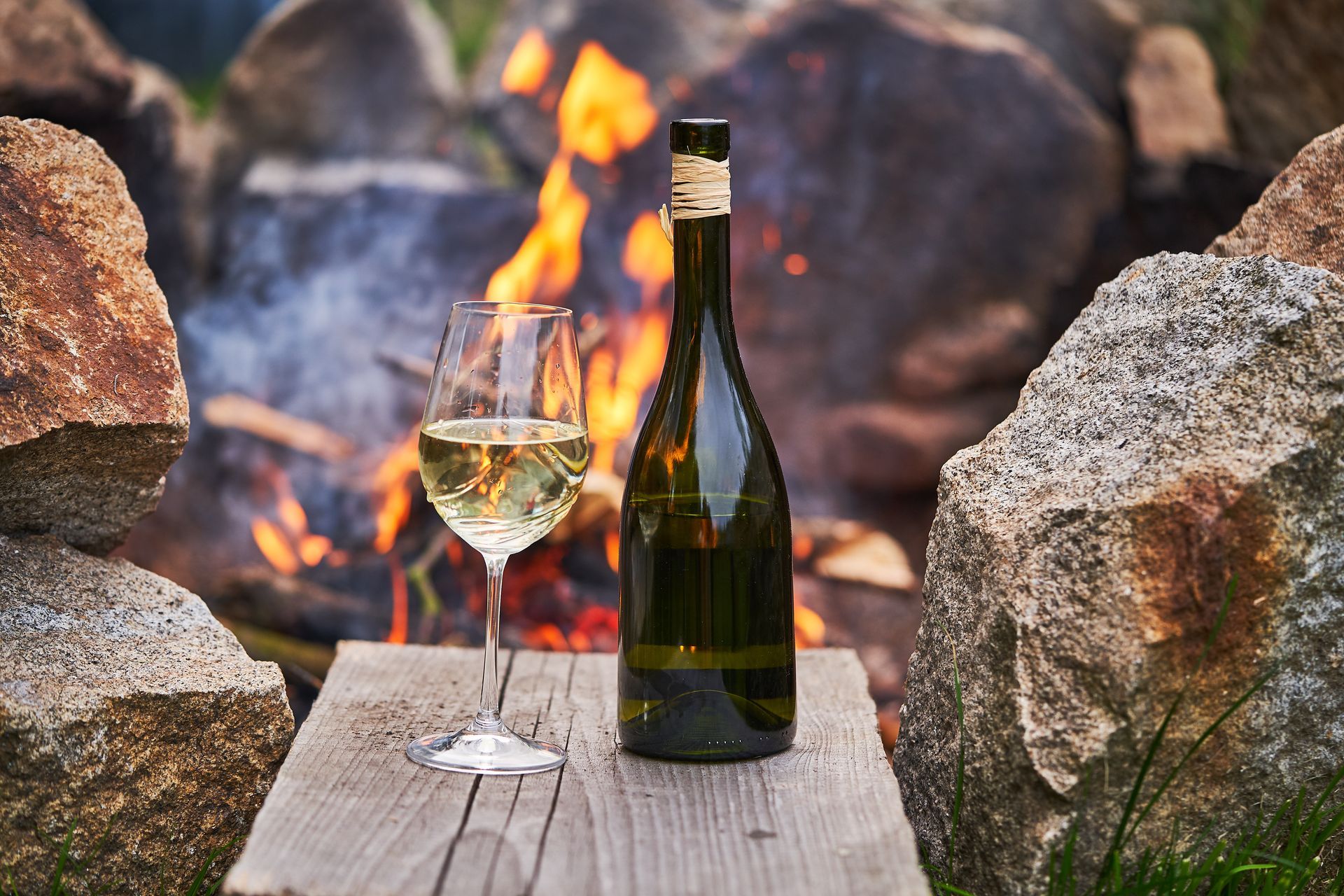 A bottle of wine and a glass of wine are on a wooden table in front of a fire pit.