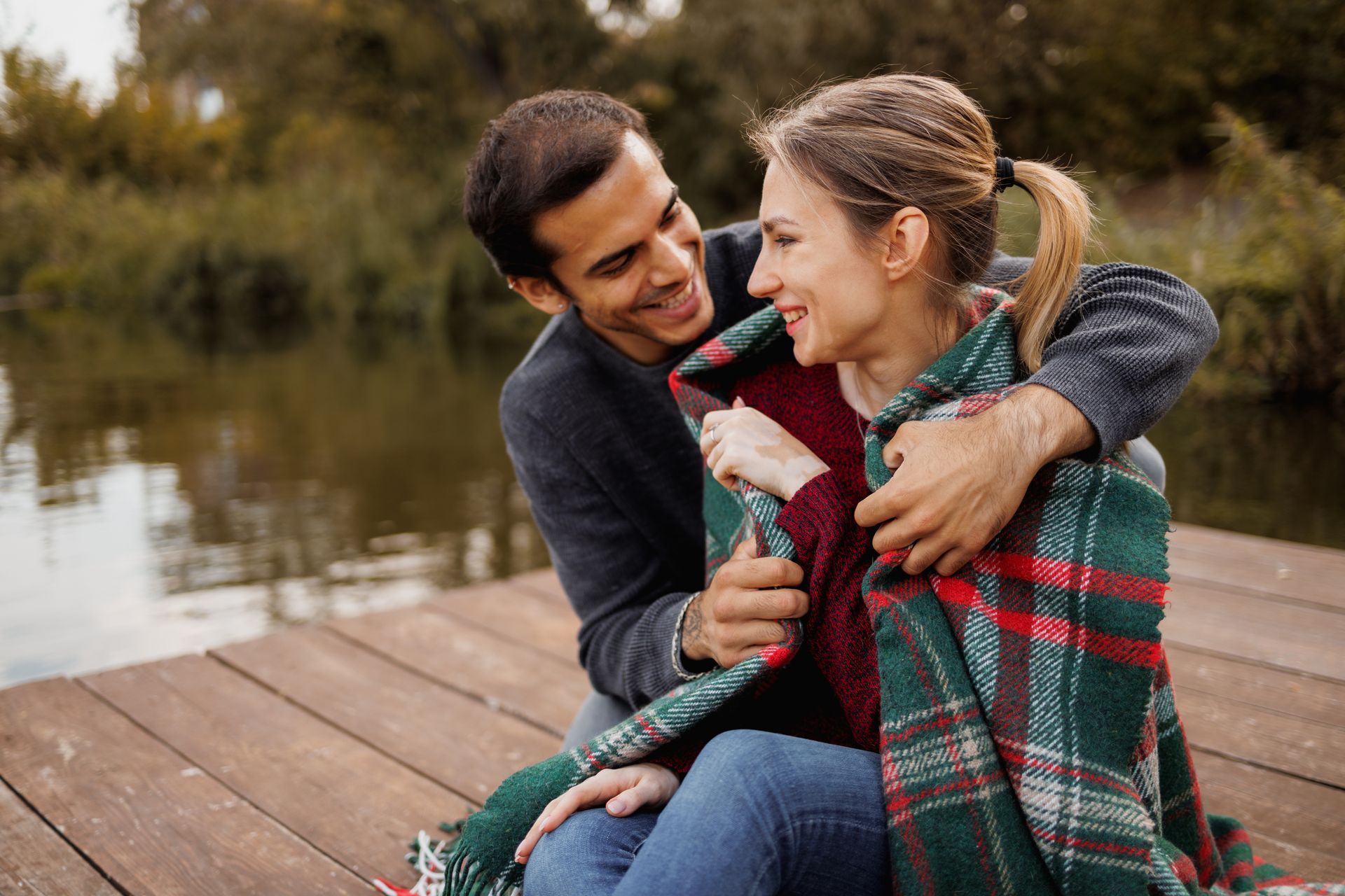 A man and a woman are sitting on a dock wrapped in a plaid blanket.