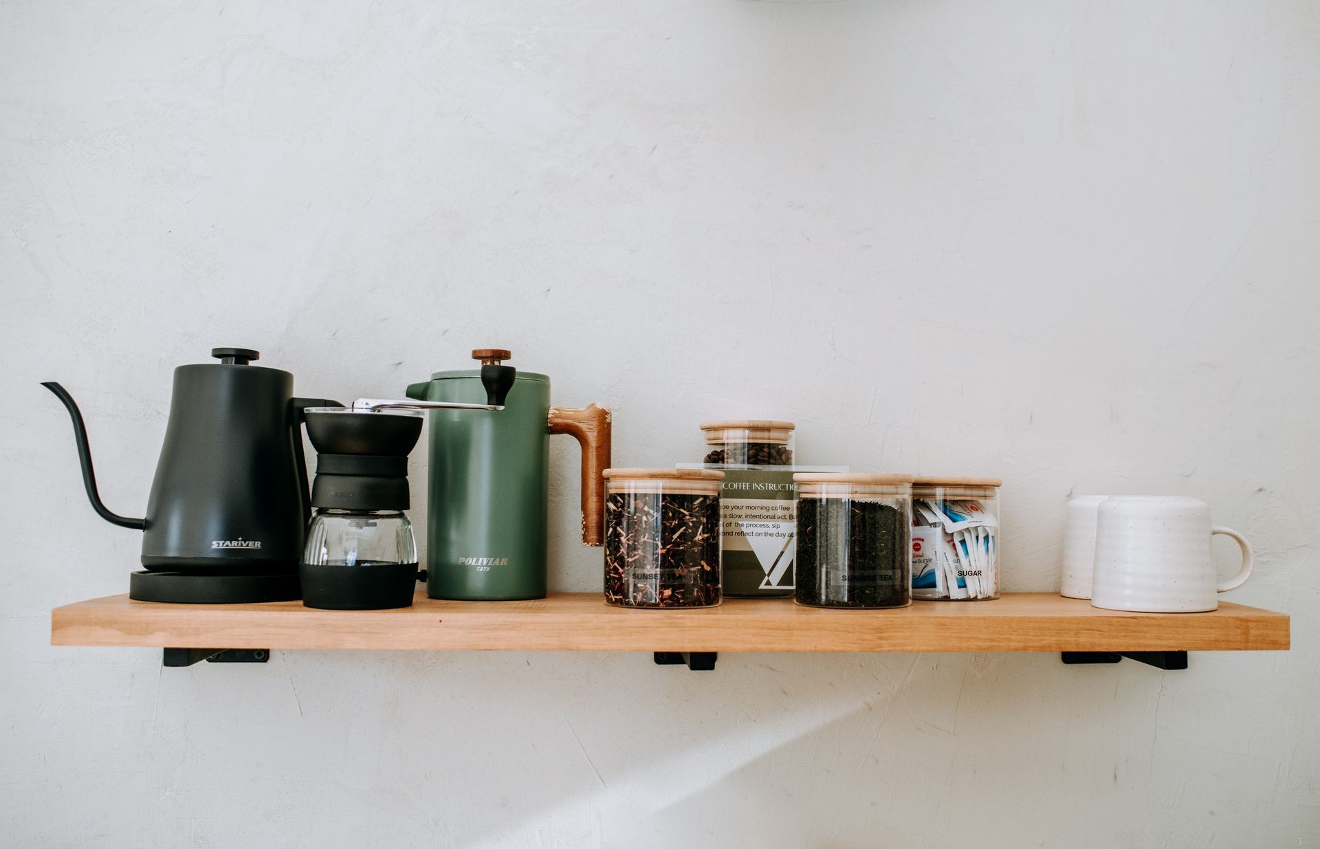 A wooden shelf with coffee pots , mugs , and jars on it.