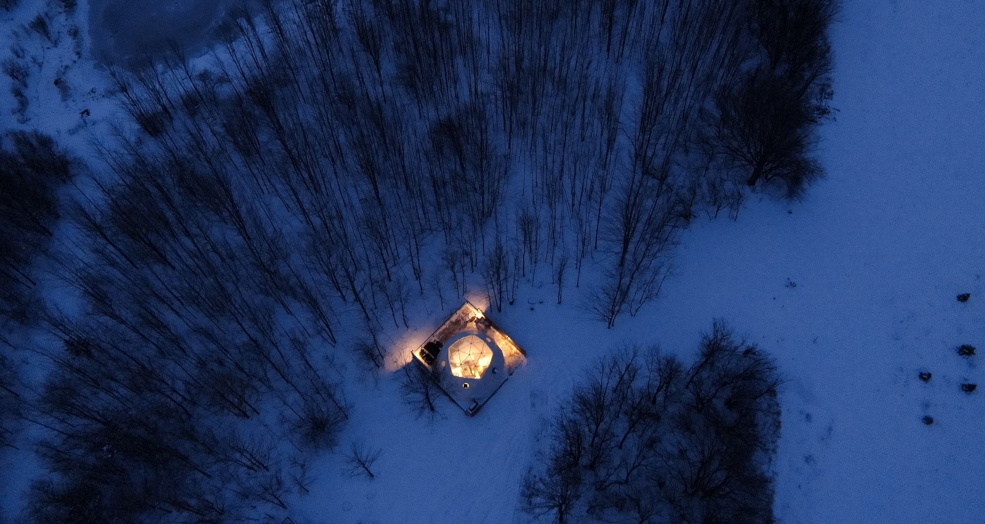 An aerial view of a small cabin at night, illuminated, surrounded by snowy trees.