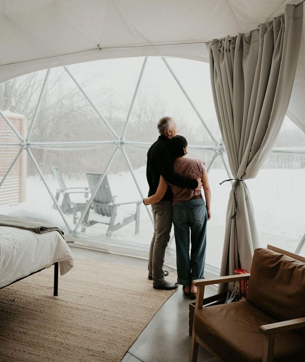 Couple embraces, looking out a dome window at a snowy landscape. Inside a glamping tent.