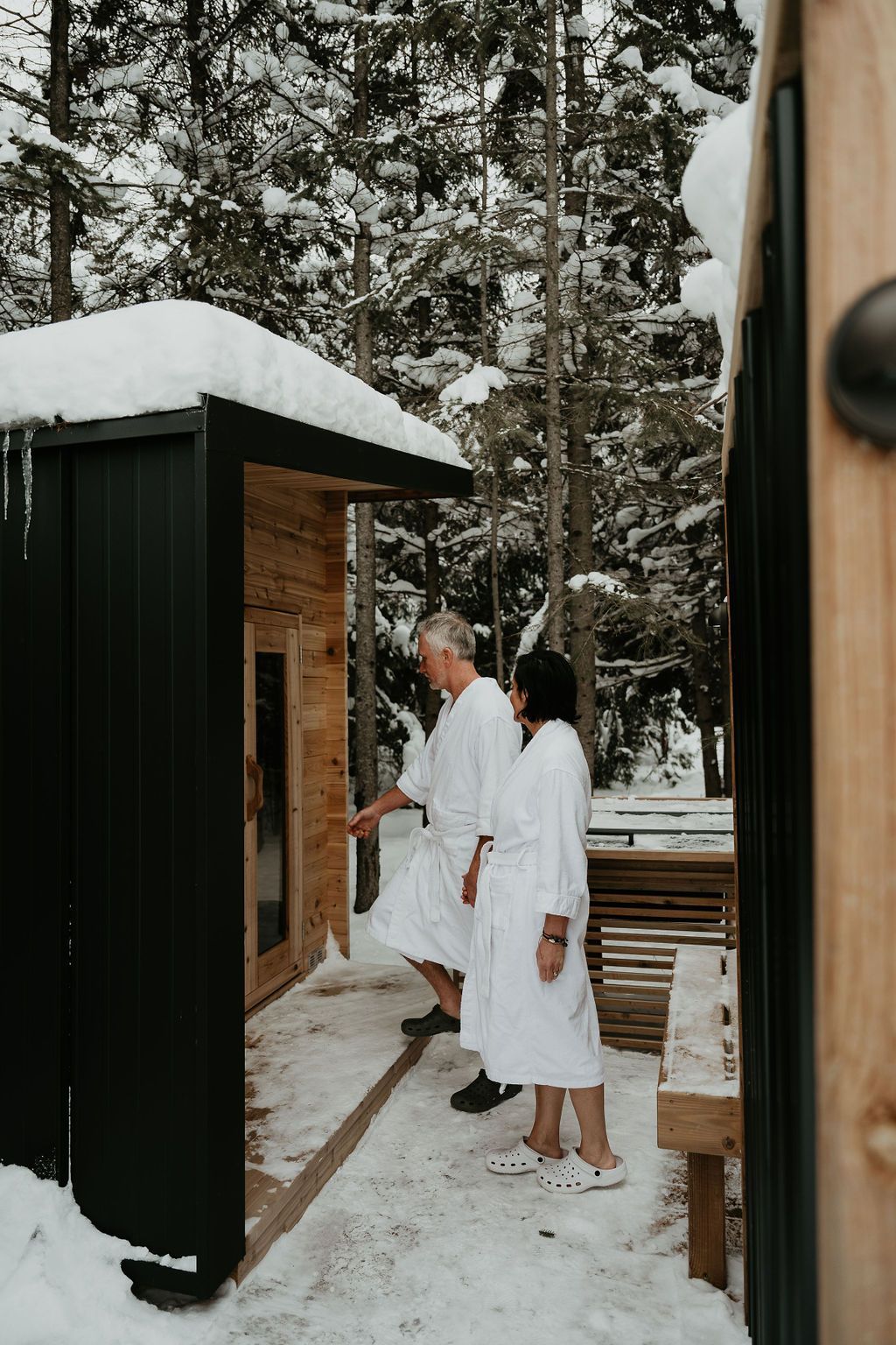 Man and woman in white robes enter outdoor sauna in snowy forest.