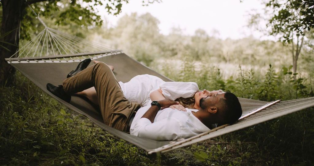 Couple relaxes in a hammock in a grassy, wooded area; man embraces woman.