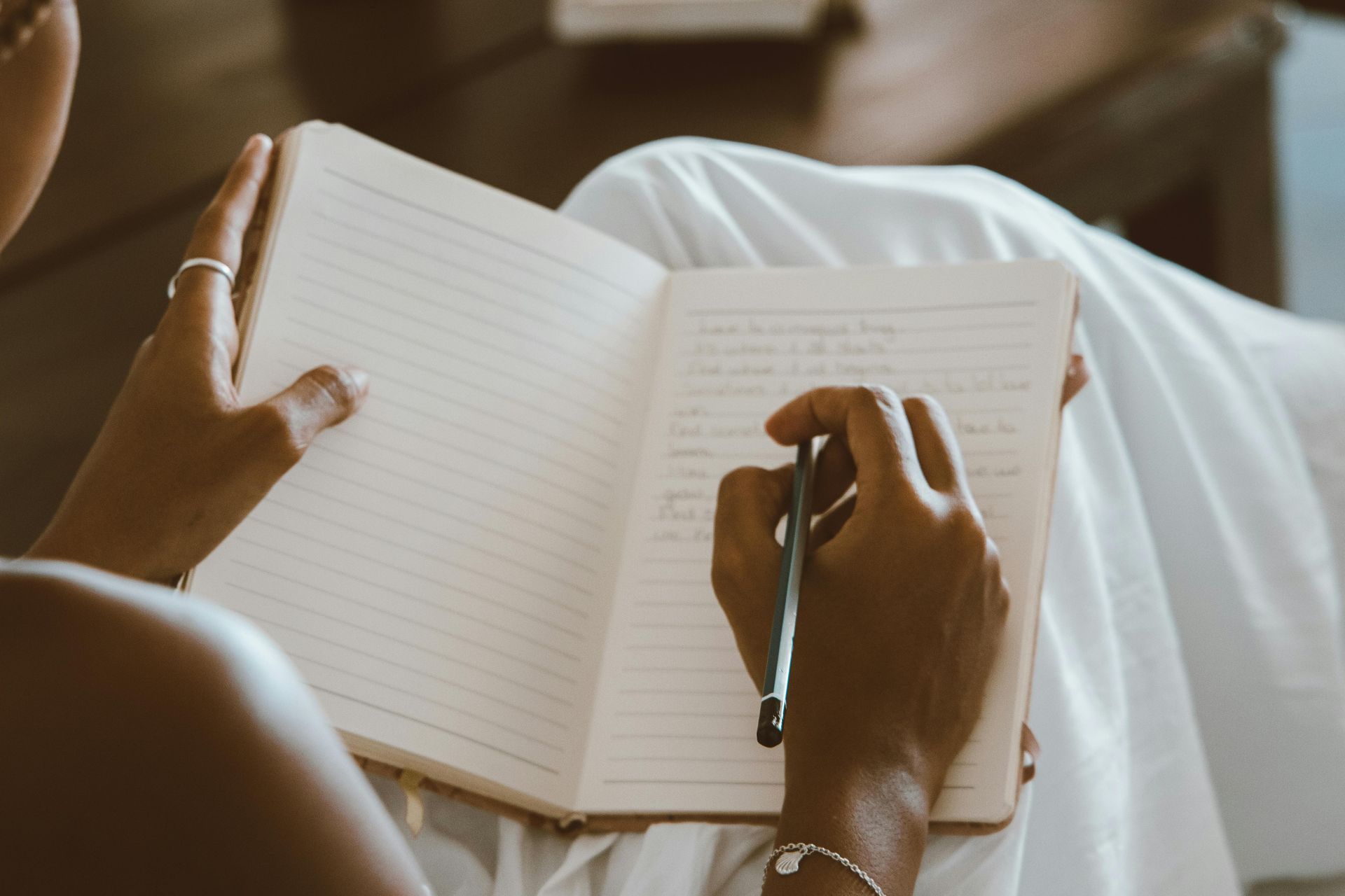 Person writing in a notebook, holding pen, sitting in white clothing.
