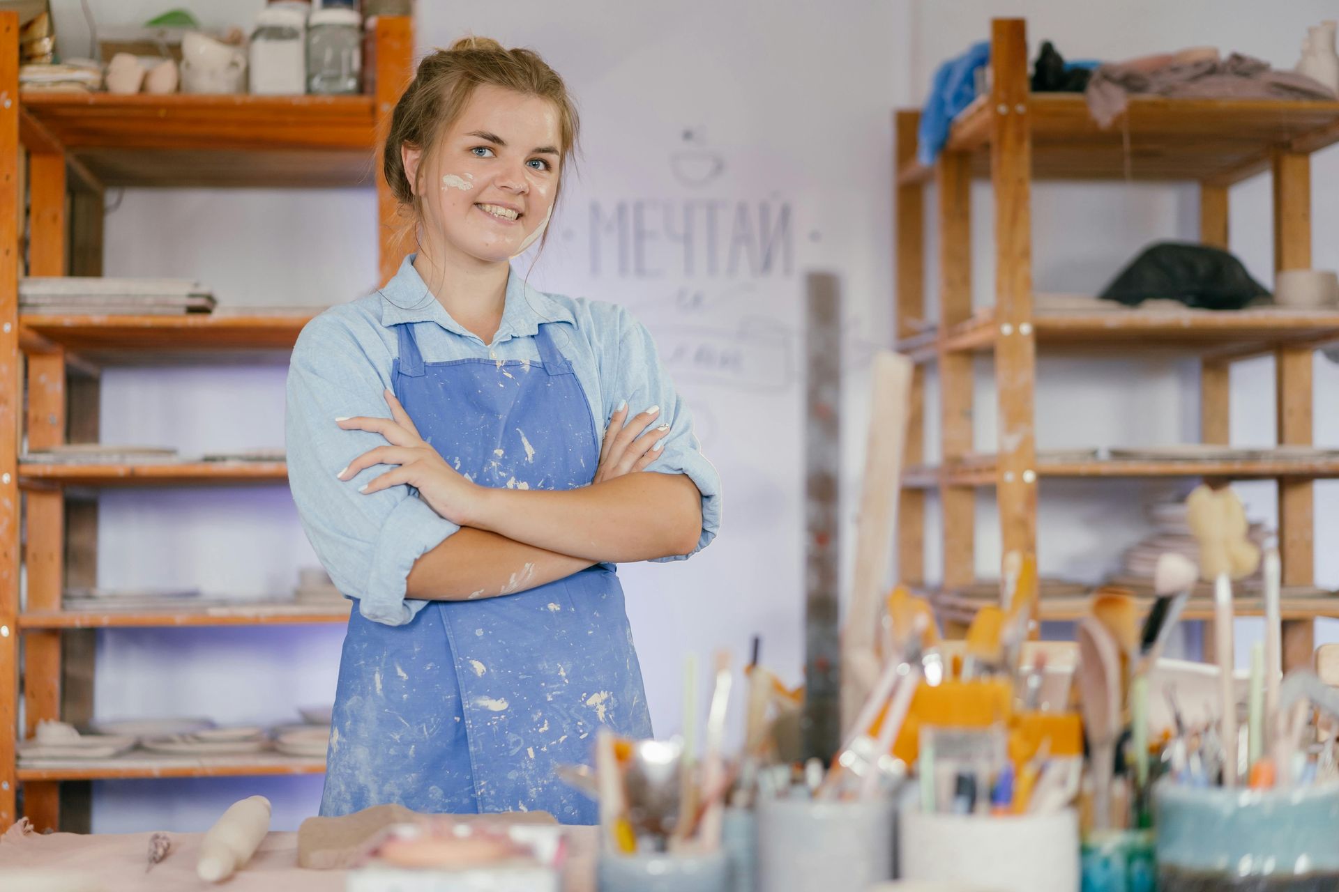 A woman in an apron is standing in a pottery studio with her arms crossed.