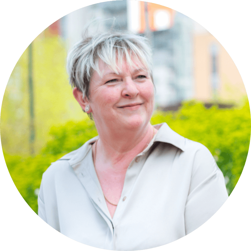 Woman with short blonde hair smiles, wearing a cream-colored collared shirt, outdoors with blurred greenery.