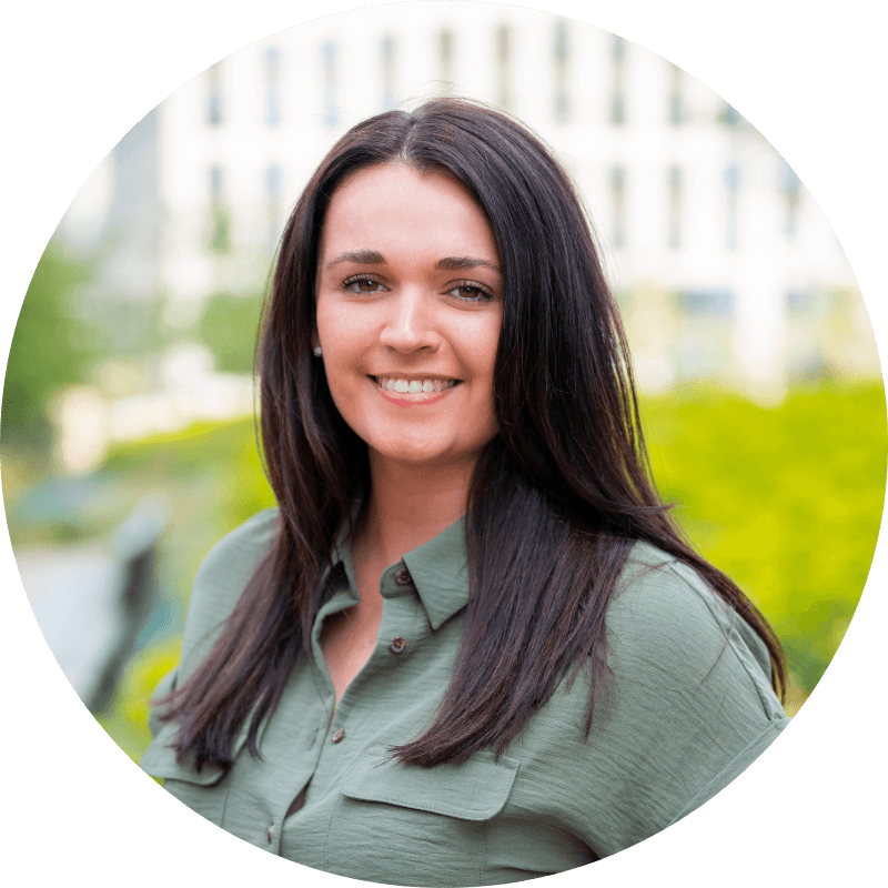 Woman with dark hair smiles, wearing a green collared shirt. Outdoors with blurred background of greenery and building.