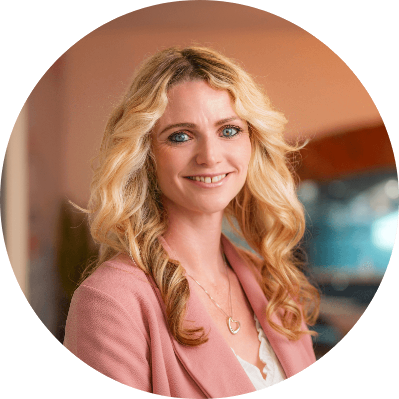 Woman with blonde curly hair smiles, wearing a pink blazer and pendant.
