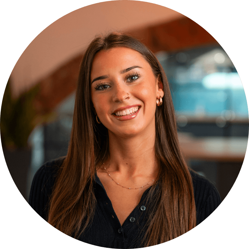 Woman with long brown hair smiles, wearing a black shirt and gold jewelry in an office setting.