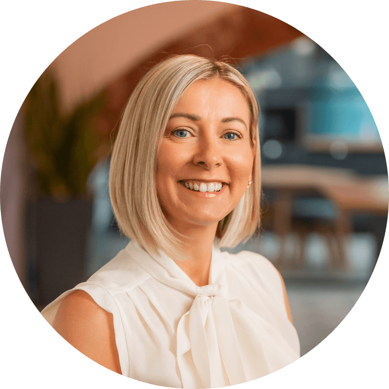 Woman with blonde hair smiling, wearing a white blouse in an office setting.