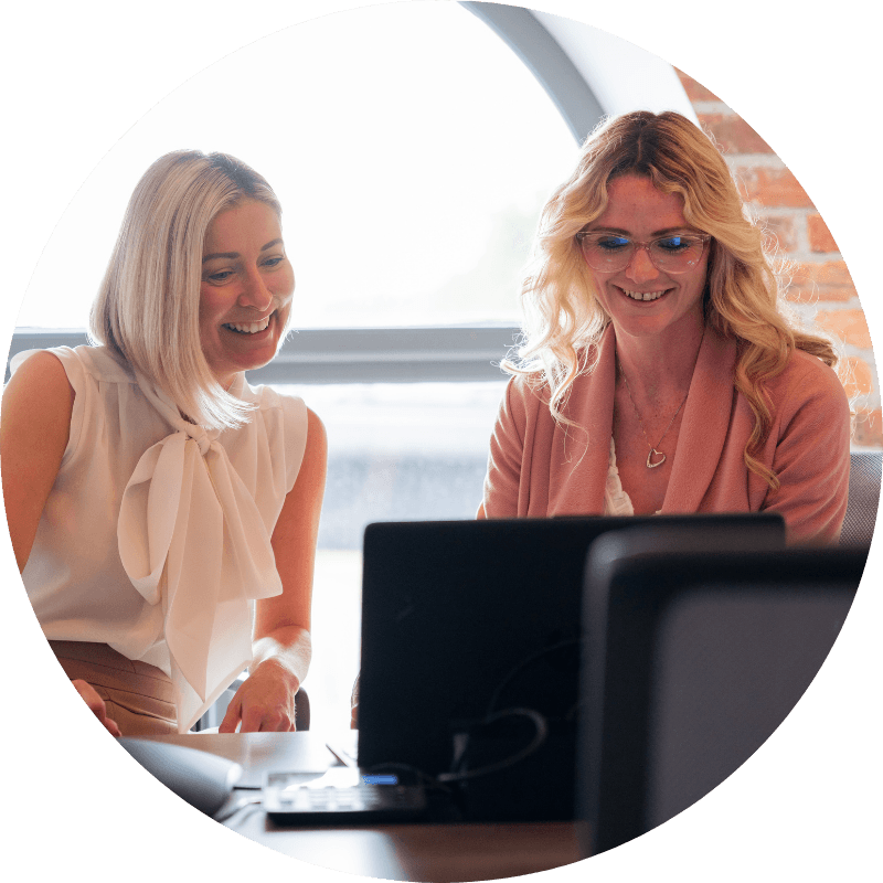 Two women smiling, looking at a laptop in an office. One in a pink jacket, the other in a cream blouse.