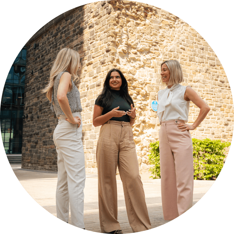 Three women talking outdoors in front of a stone building; one gesturing.