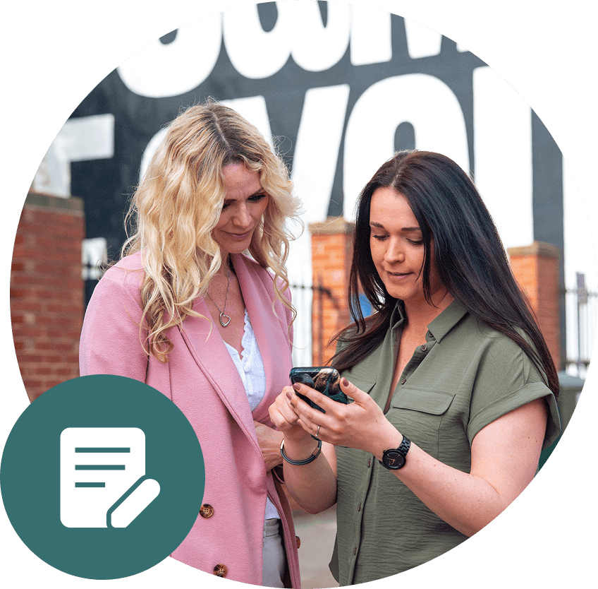 Two women looking at a phone outdoors in front of a sign. One wears pink, the other a green shirt.