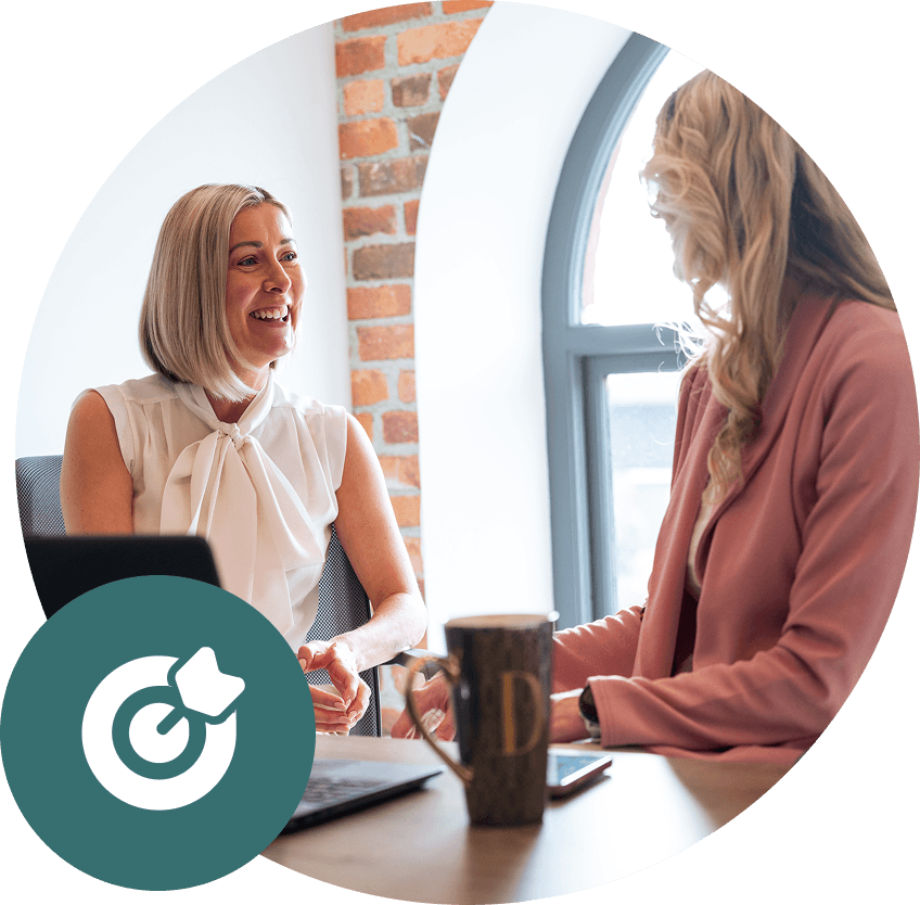 Two women smiling, having a conversation at a table with a laptop, coffee mug, and brick wall background.