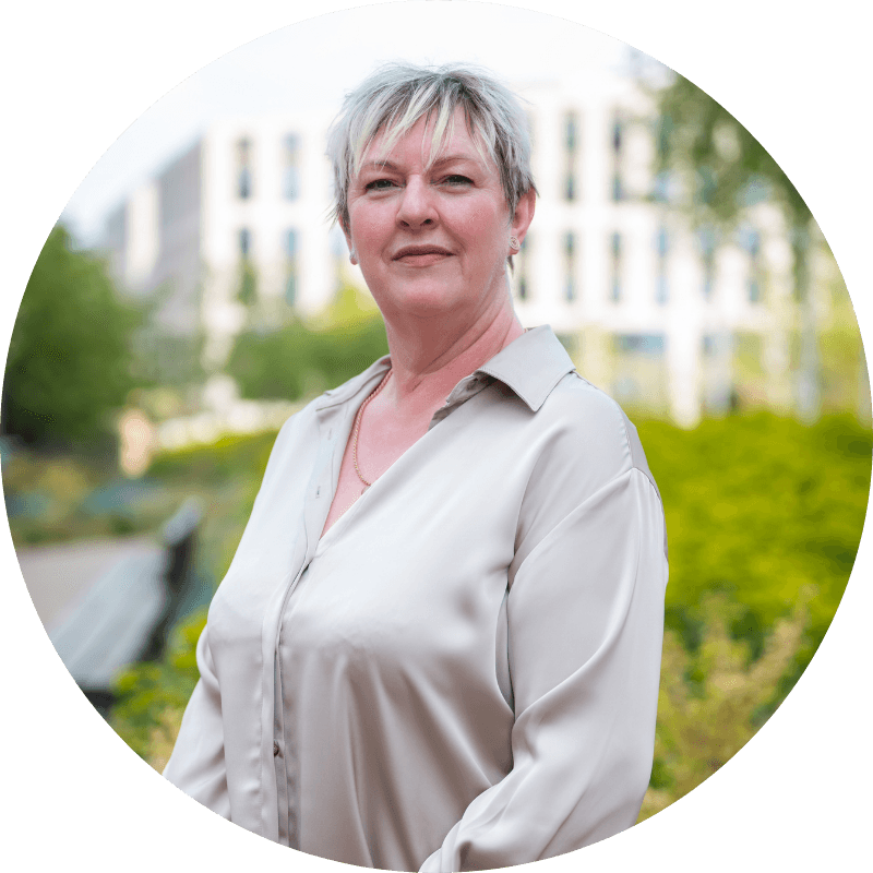 Woman with short grey hair, wearing a cream-colored shirt, smiling. Outdoors, building in background.