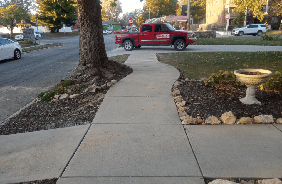 A concrete sidewalk curves around a tree, passing a red pickup truck on a street.