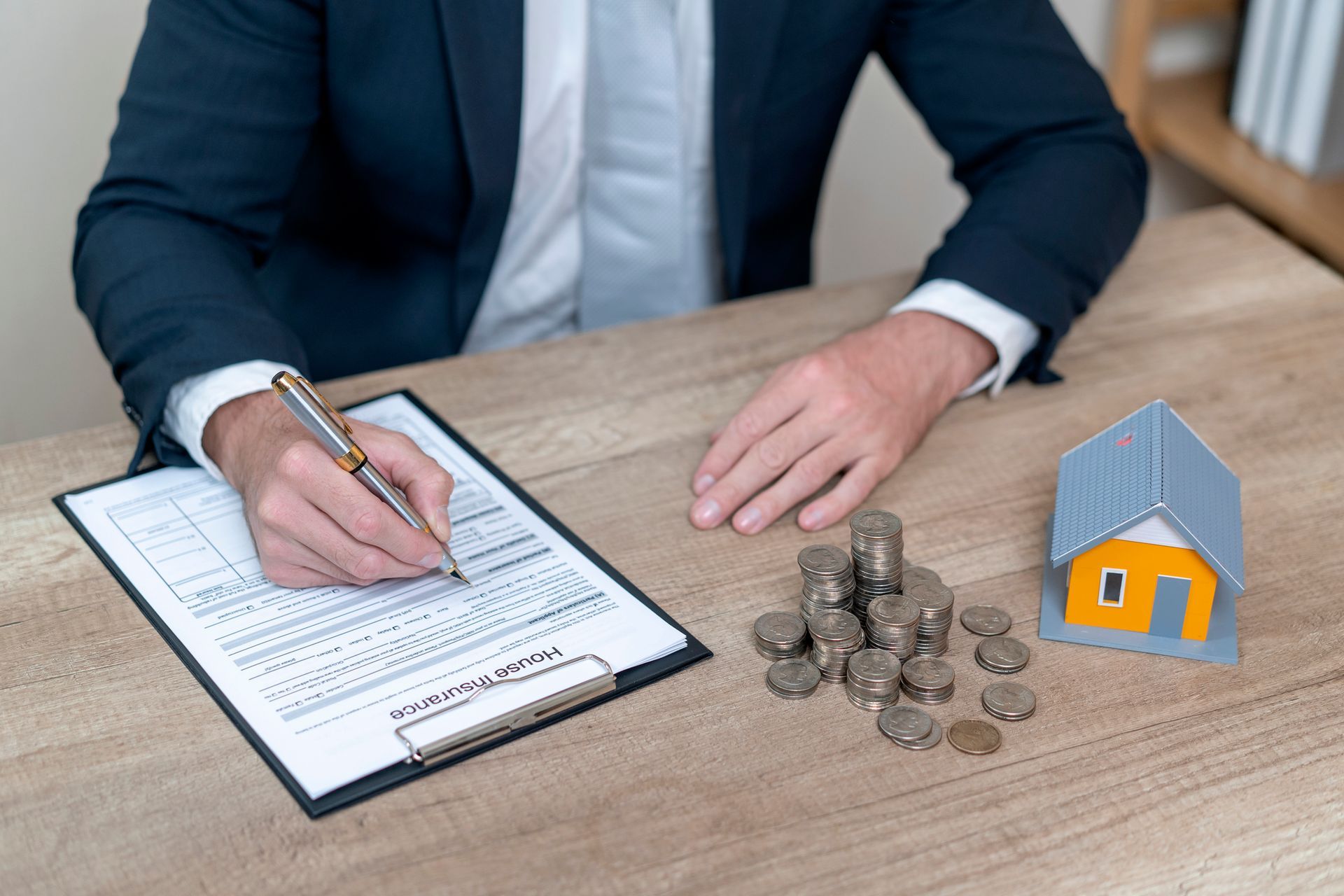 Man in suit filling out paperwork next to a pile of coins and a toy house on a table.