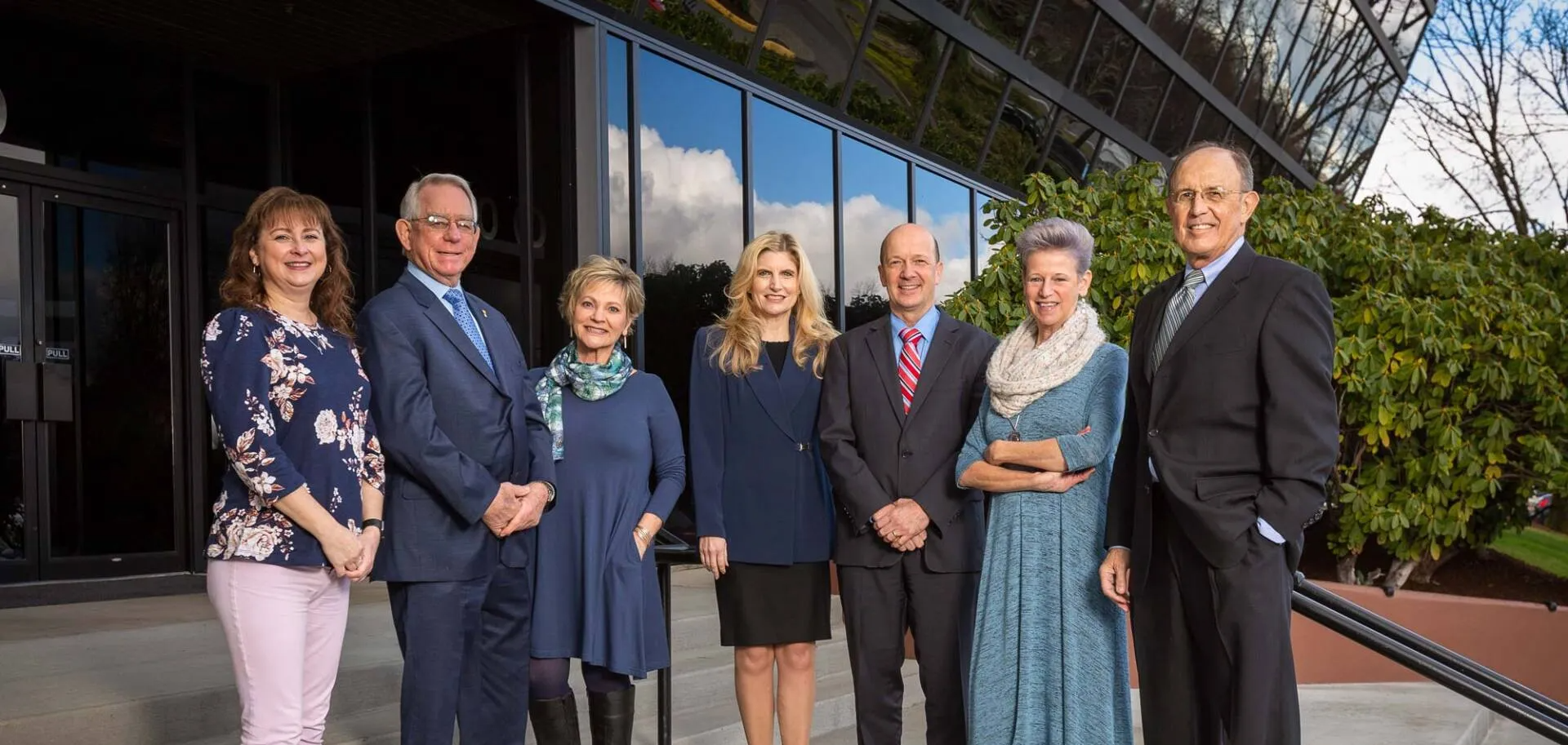 Group of people standing in front of a modern building. The people are well-dressed, and the setting appears to be daytime.